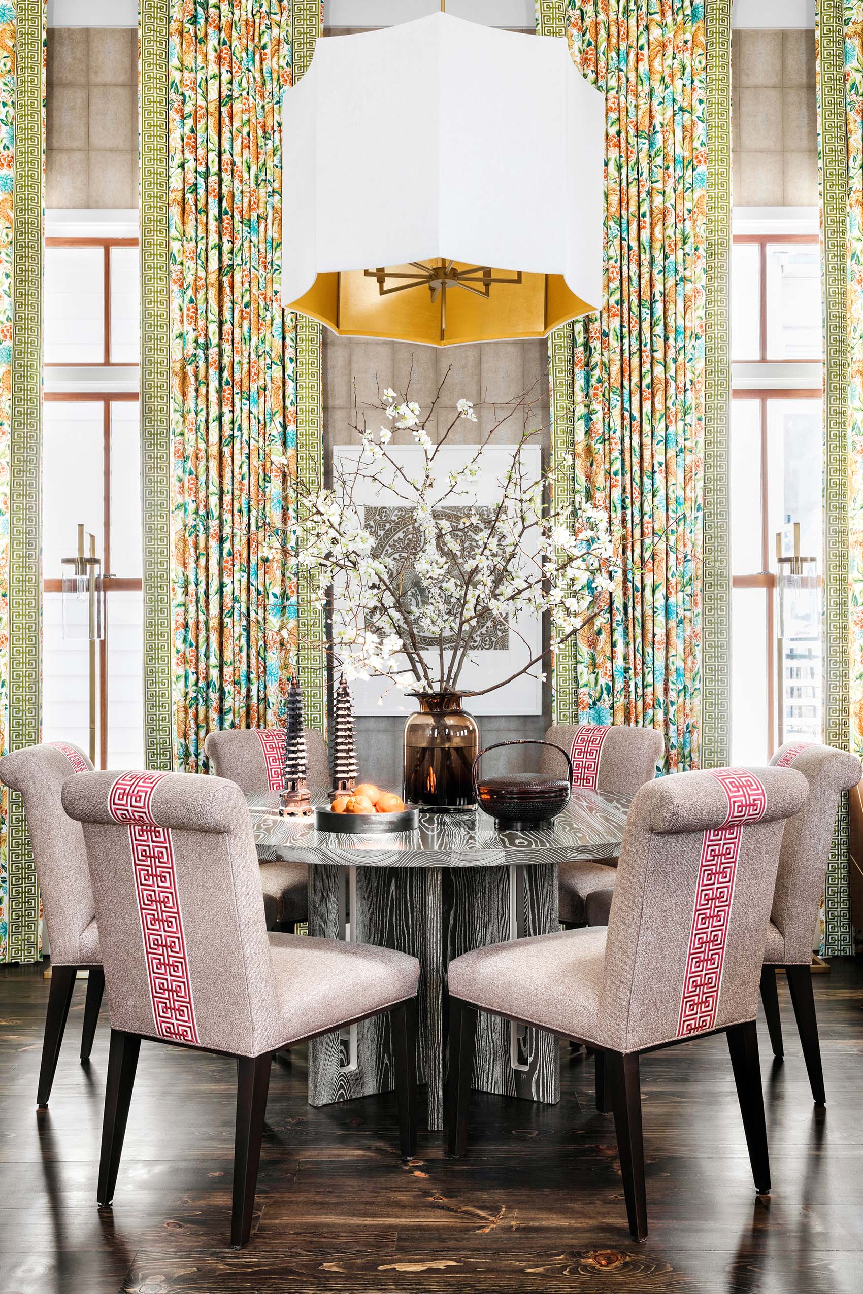 A formal dining room featuring a circular wood grain table, upholstered chairs with red Greek key trim, and tall floral patterned curtains.