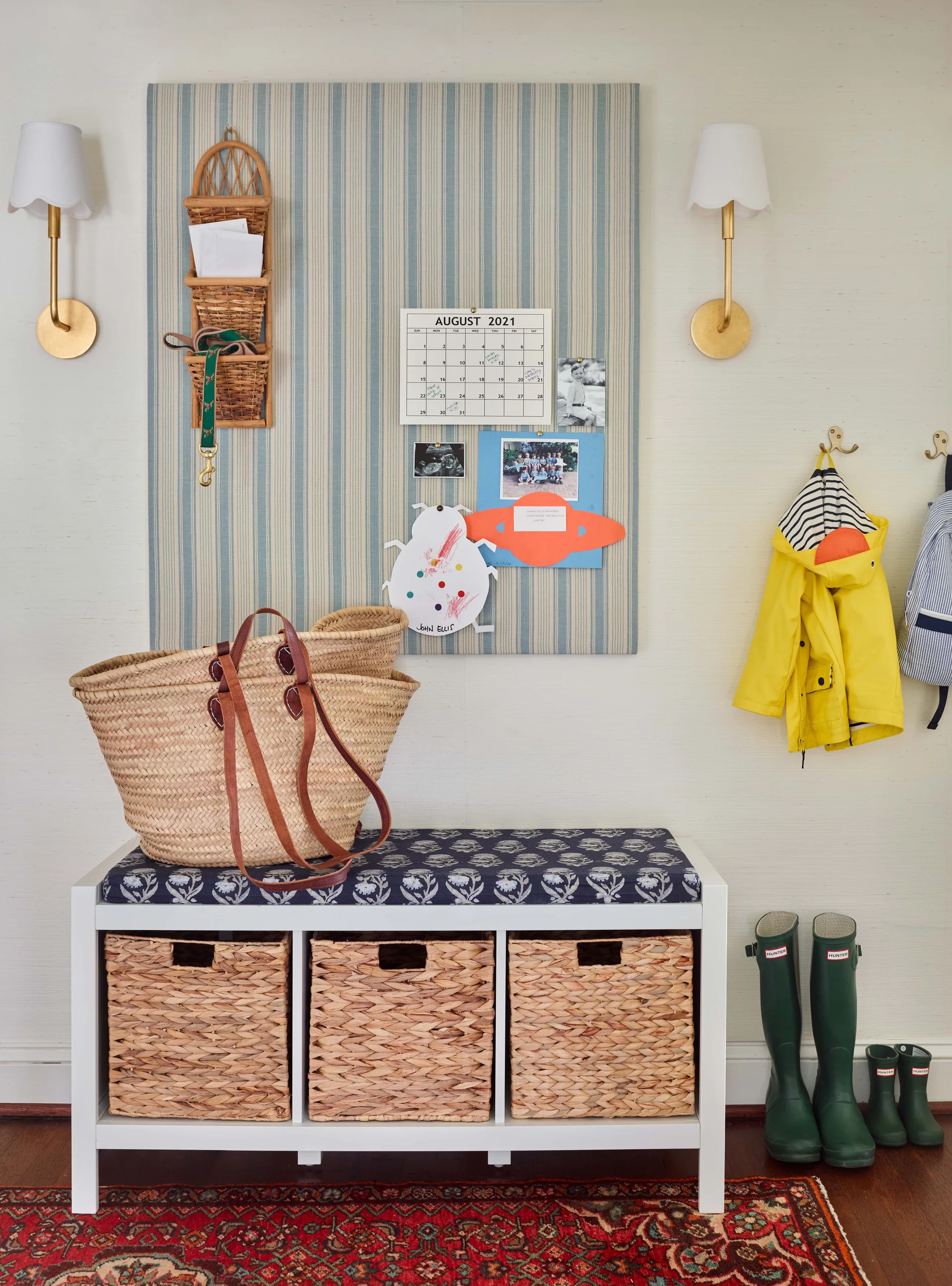 A white mudroom storage bench with woven baskets sits beneath a blue striped fabric pinboard flanked by brass wall sconces.