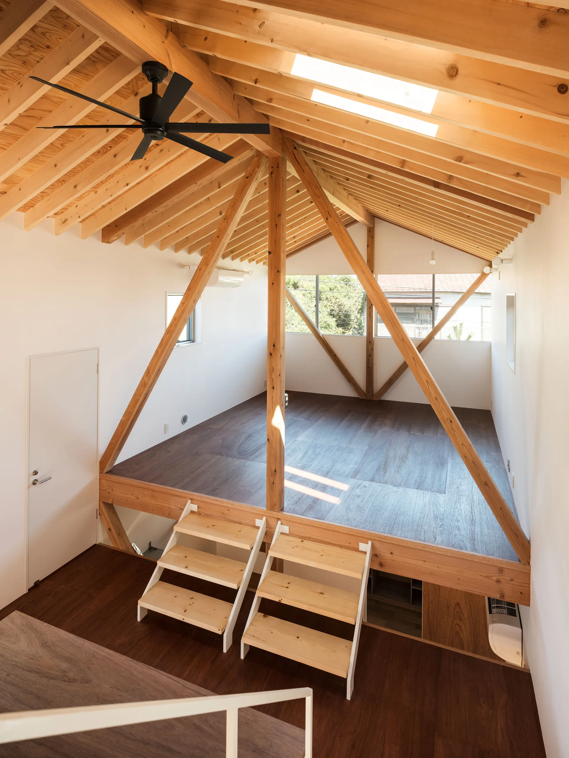A bright lofted interior featuring exposed wooden ceiling beams, white walls, a black ceiling fan, and two short wooden ladders.