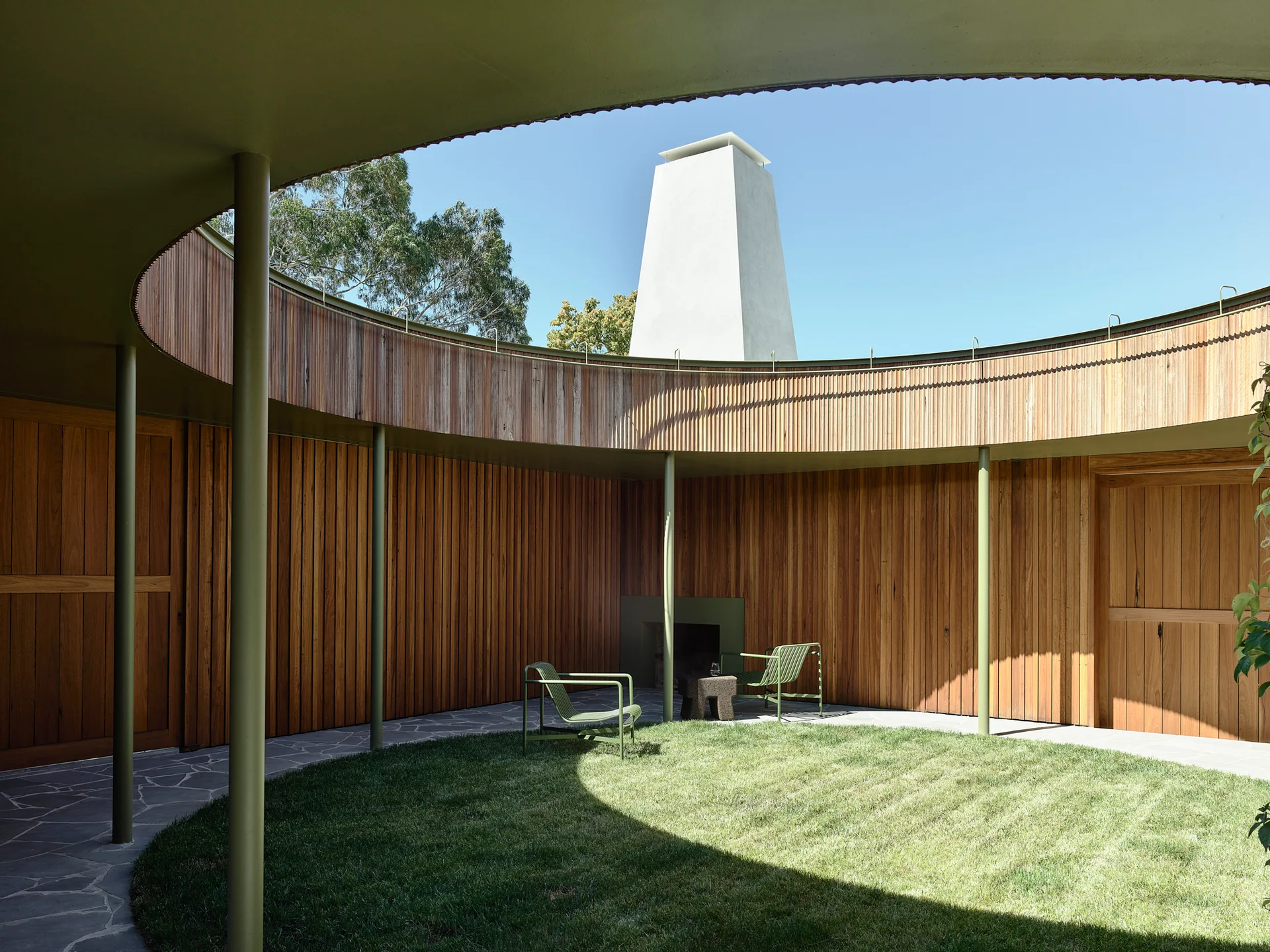 A circular outdoor courtyard with curved vertical wood-paneled walls, a flagstone patio bordering a round lawn, and green structural columns.