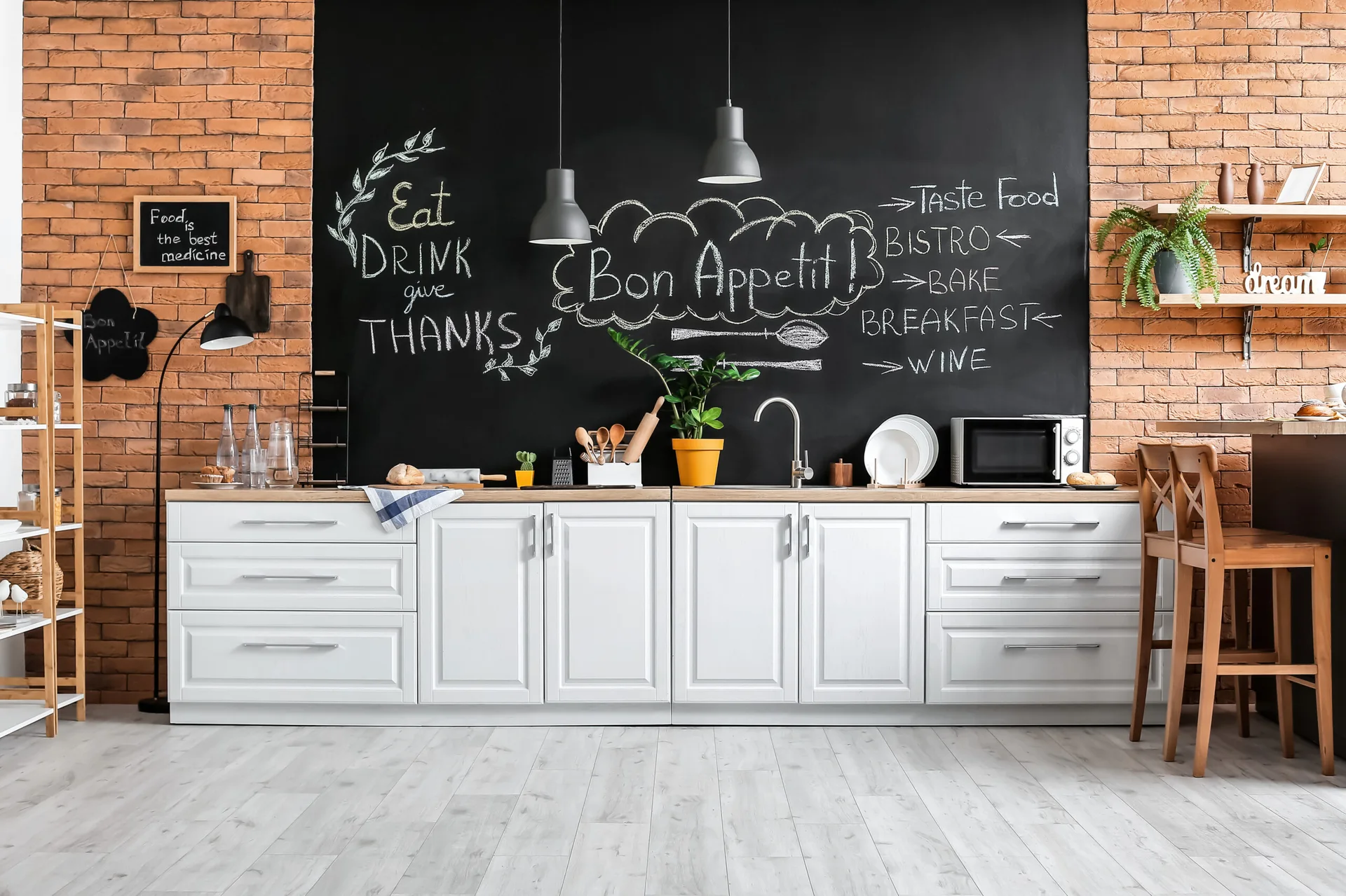 White shaker-style kitchen cabinets with light wood countertops set against a large black chalkboard accent wall flanked by exposed brick.