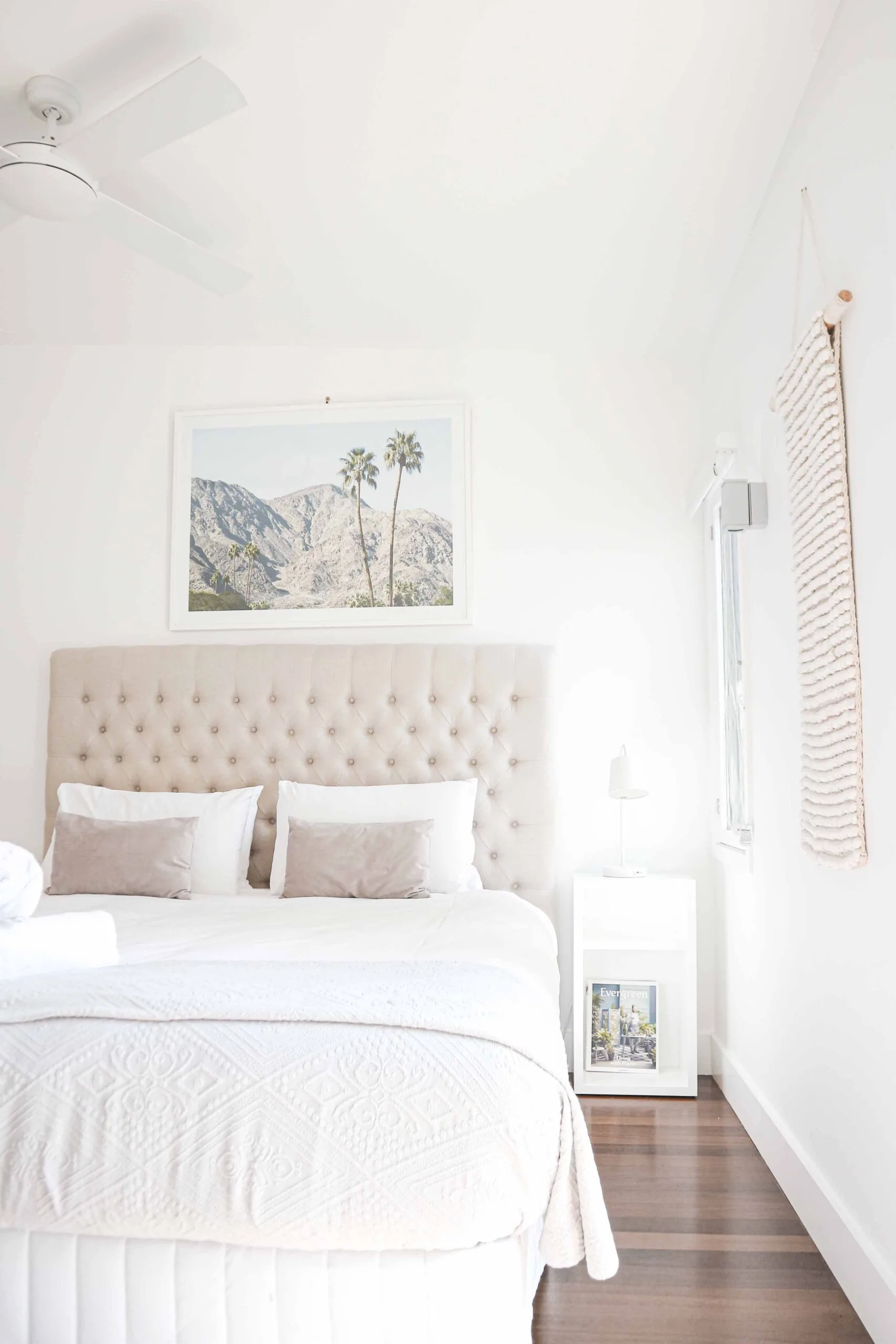 A bright white bedroom featuring a tufted beige headboard, white bedding with taupe accent pillows, and a framed desert landscape print on the wall.