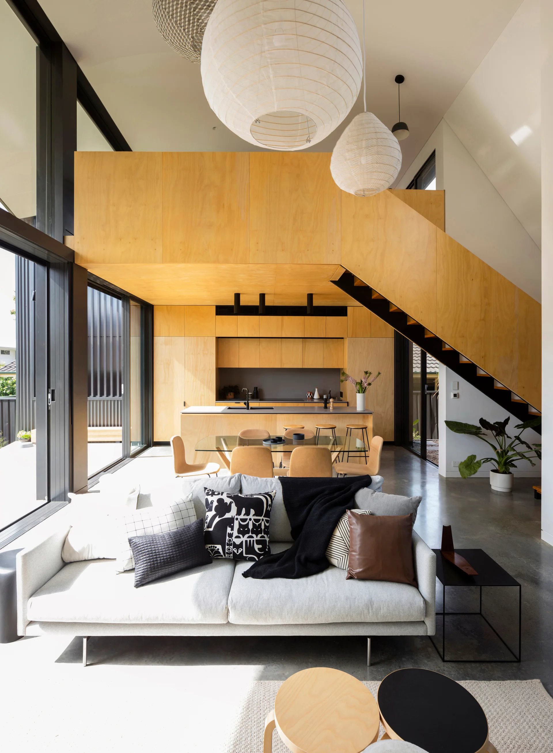 A modern open-concept living room features a light gray sofa, polished concrete flooring, a plywood loft structure, and hanging paper pendant lights.