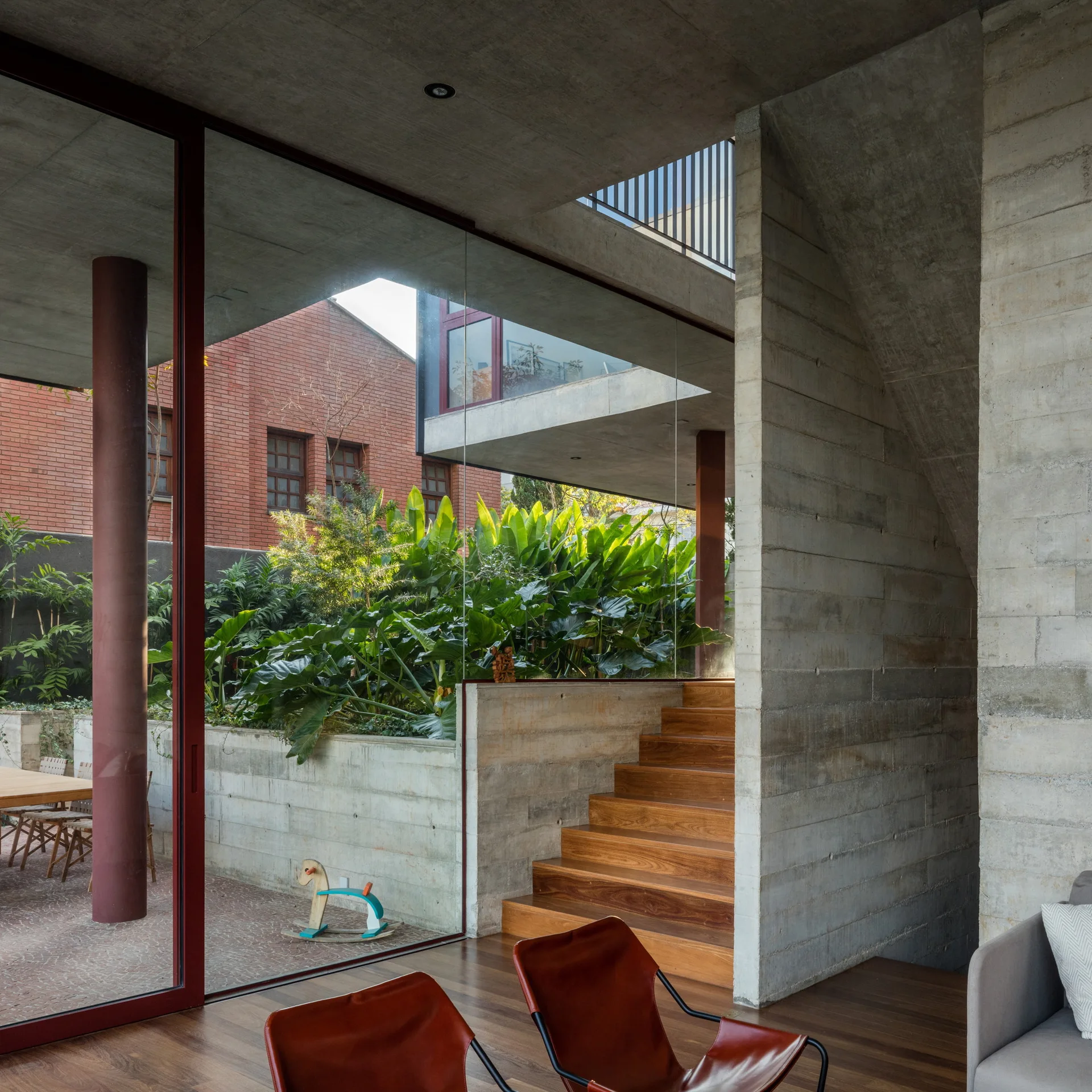 A modern interior featuring board-formed concrete walls, a dark wood staircase, and a large glass partition showing an outdoor garden patio.