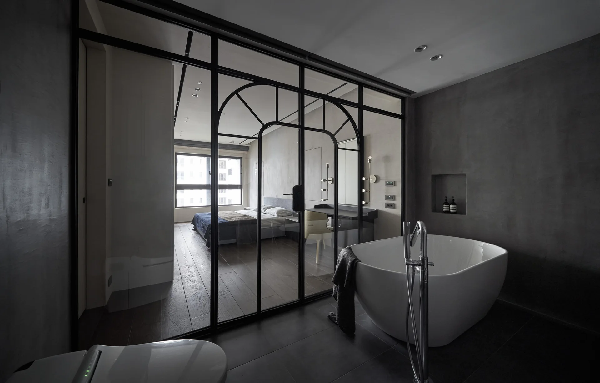 A modern gray bathroom with a freestanding white bathtub looking through an arched black metal glass partition into an adjacent bedroom.