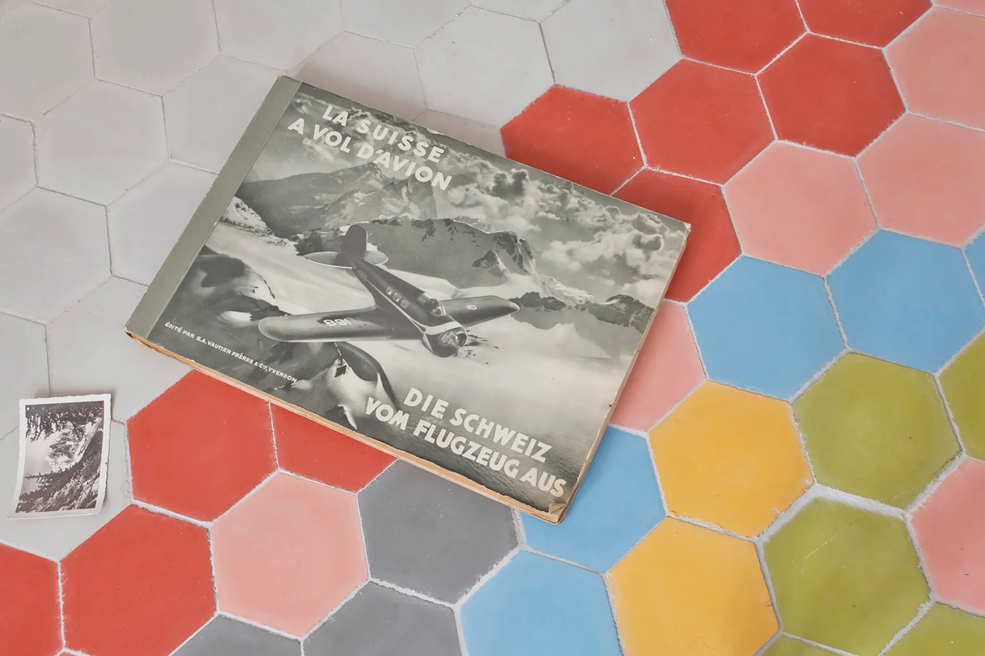 A close-up view of multicolored matte hexagonal floor tiles with a vintage book and a small black and white photograph resting on them.