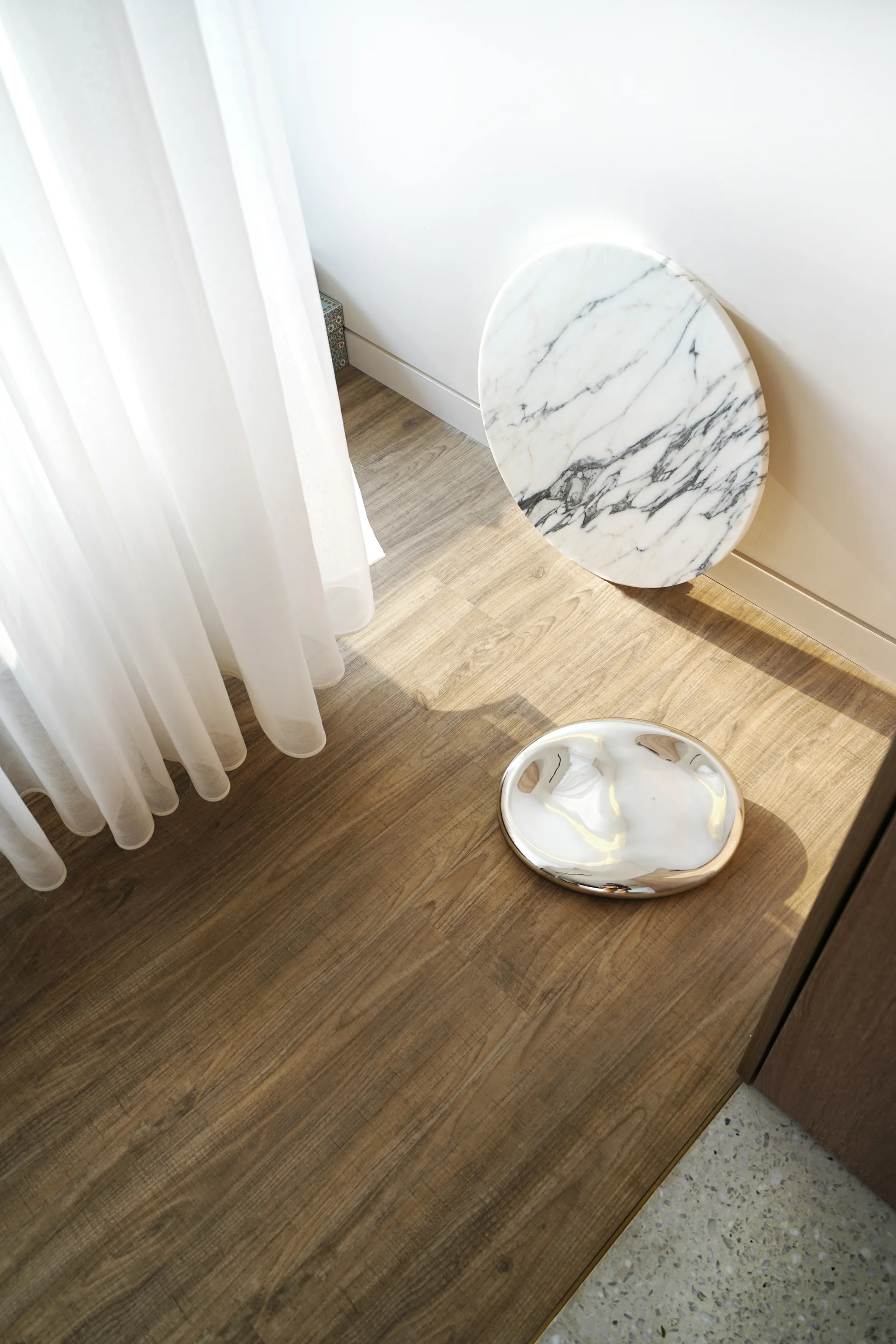A circular white marble slab leans against a wall next to a polished silver dish on a light brown wood floor beside a sheer white curtain.