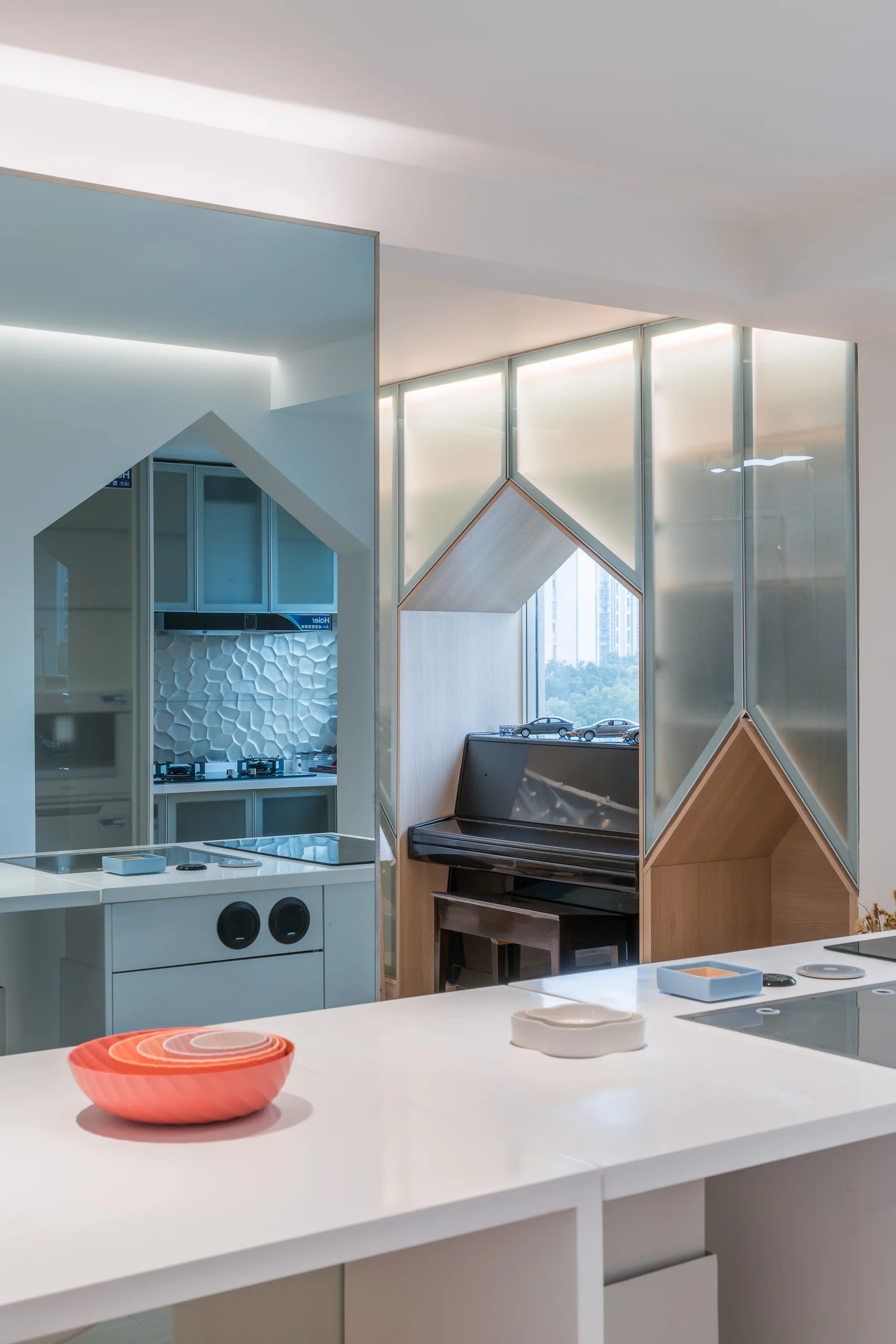 A white kitchen island sits in the foreground, with a geometric frosted glass and light wood partition framing a black piano in the background.