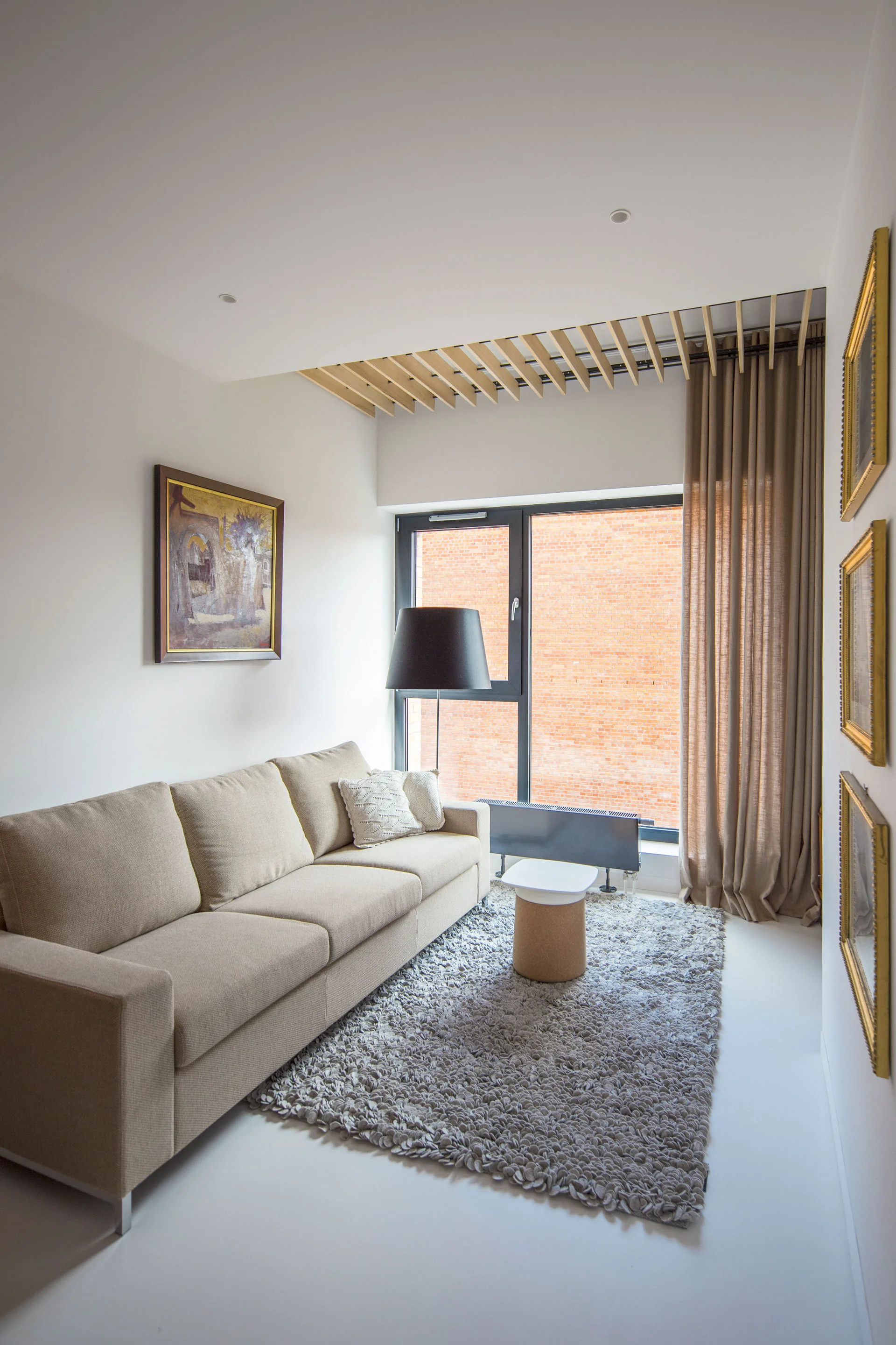 A neutral modern living room featuring a beige sofa on a highly textured grey rug next to a window with wooden ceiling slats.