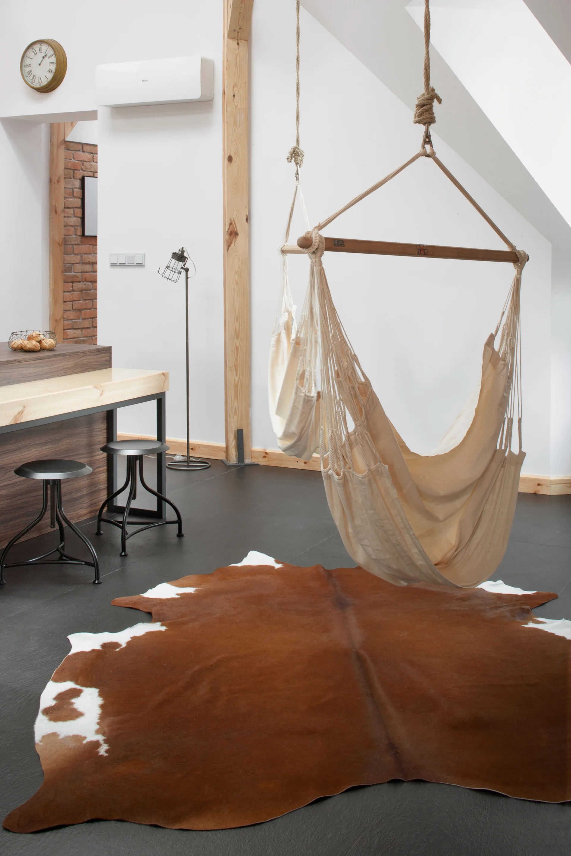 A white canvas hammock chair hangs from the ceiling above a brown and white cowhide rug in a living room with dark flooring and bar stools.