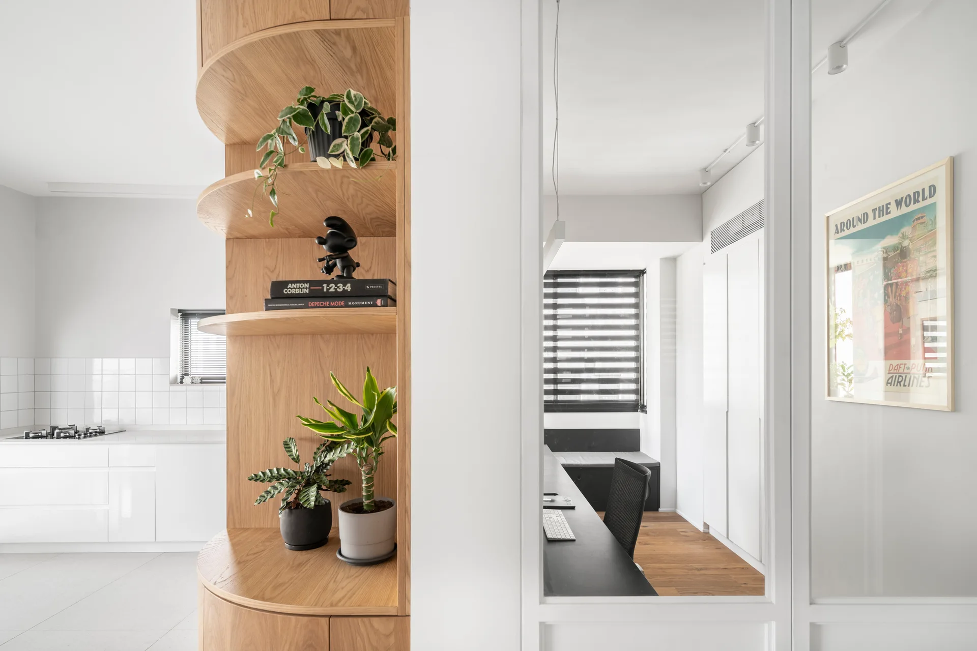 A curved oak shelving unit holding plants stands next to a white-framed glass partition looking into a home office with a desk.
