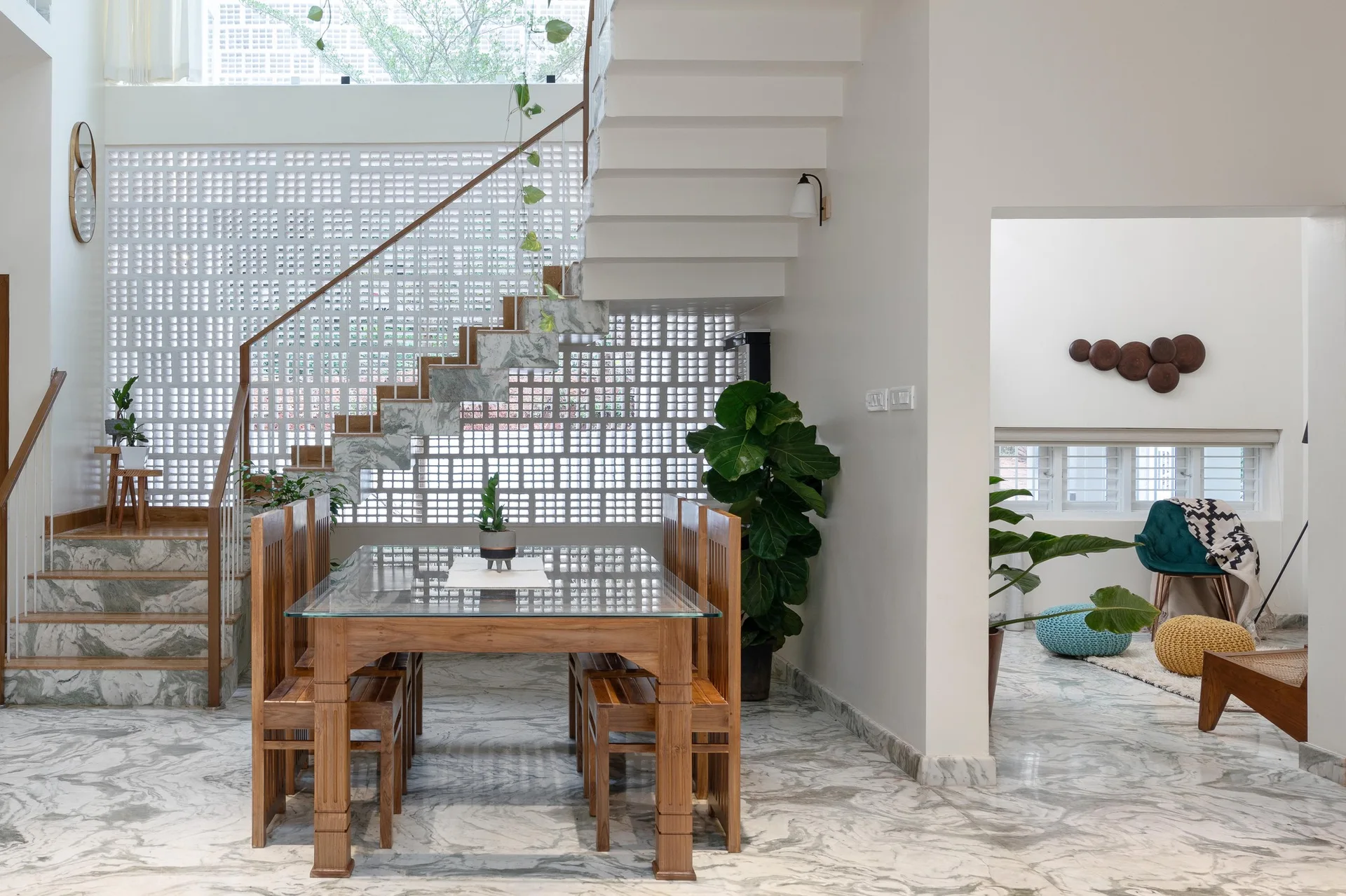 A modern dining room features a glass-topped wooden table, a marble-clad staircase, and a white breeze block accent wall.