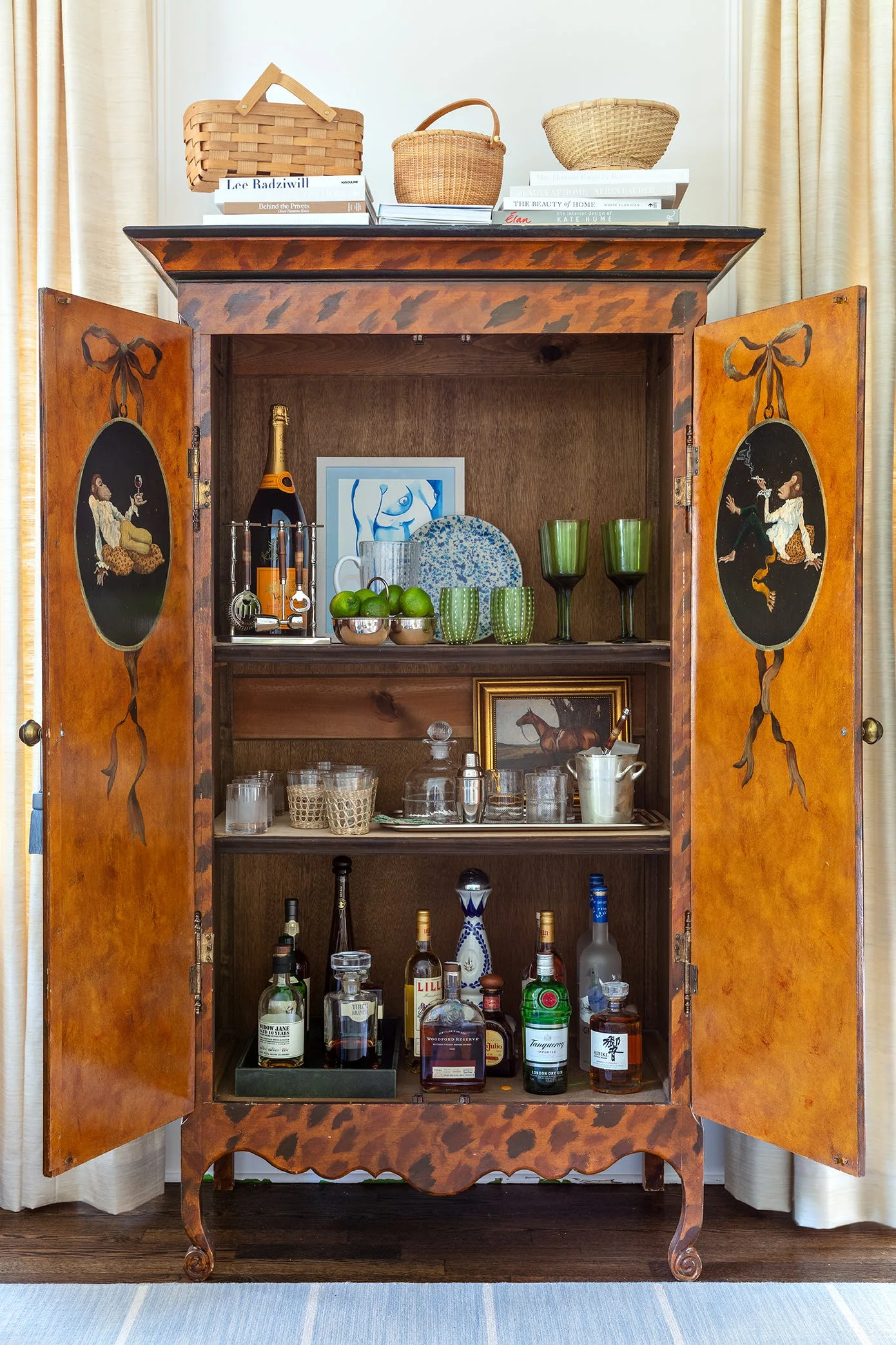 An open tortoiseshell-patterned wooden cabinet styled as a home bar, stocked with glassware and liquor bottles beneath stacked baskets.