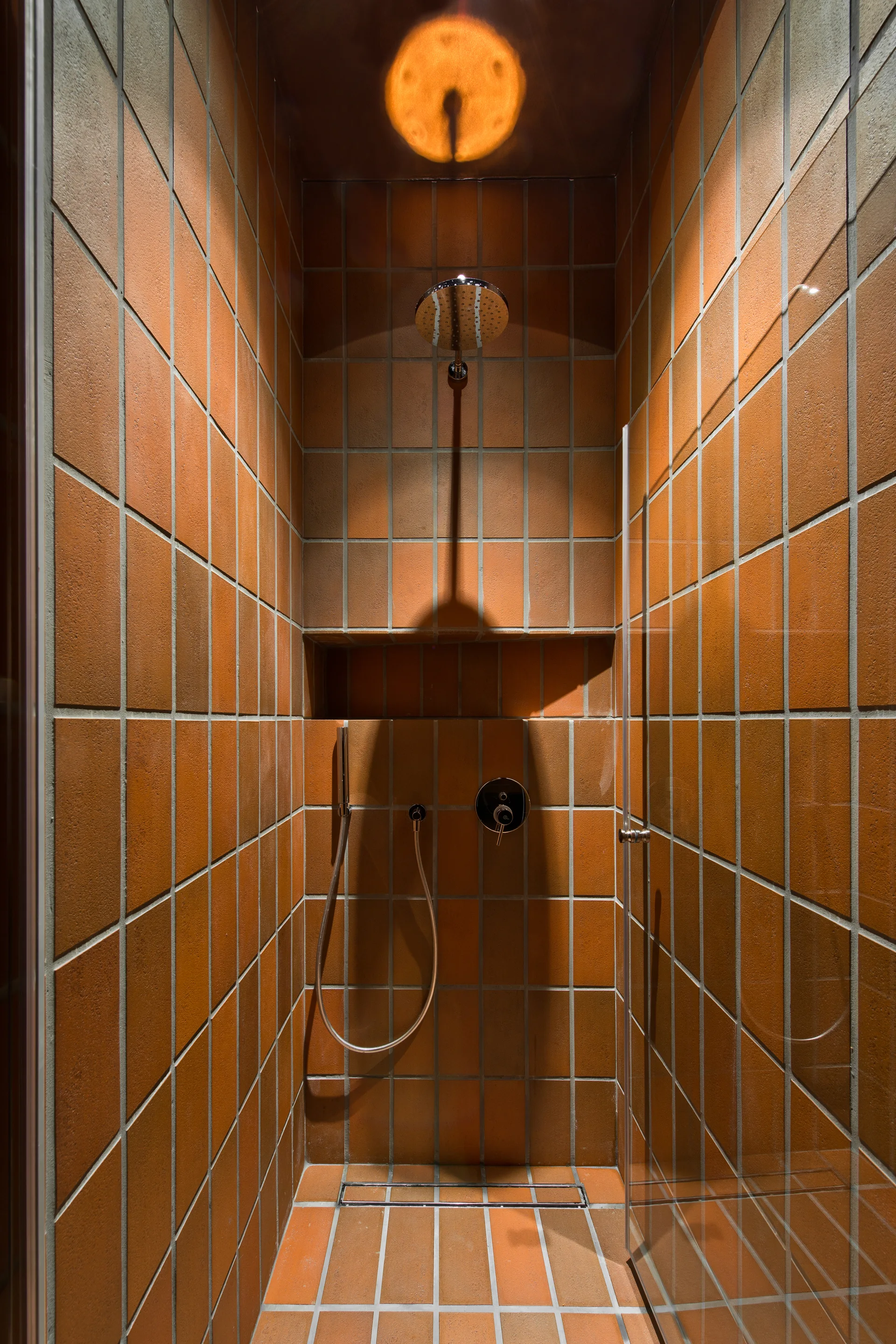 A walk-in shower stall lined with square terracotta tiles, featuring chrome fixtures, a recessed storage shelf, and a glass door.
