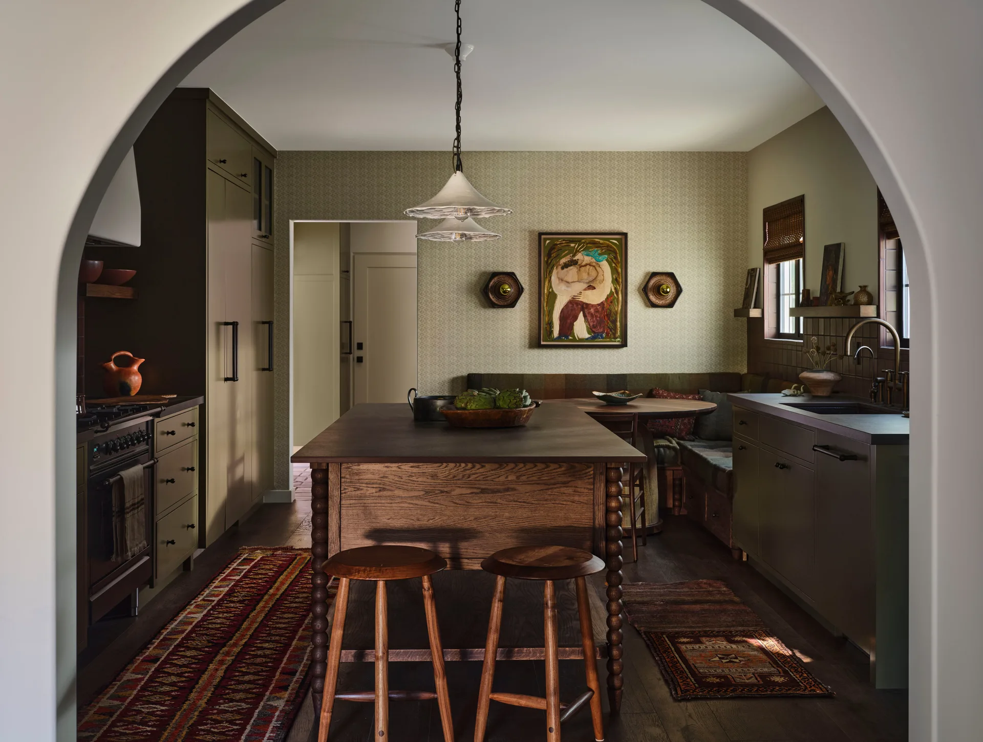 A view through a plaster archway shows an olive green kitchen with a wooden island, wooden stools, and a cozy corner breakfast banquette.