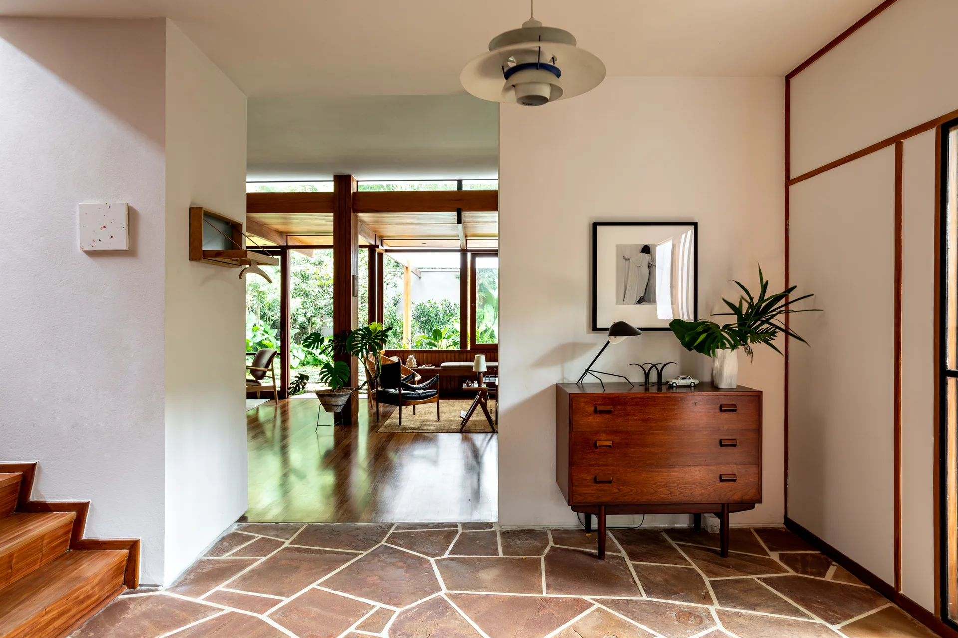 A mid-century modern entryway features flagstone flooring, a wooden chest of drawers, and a staircase leading to an open living area.