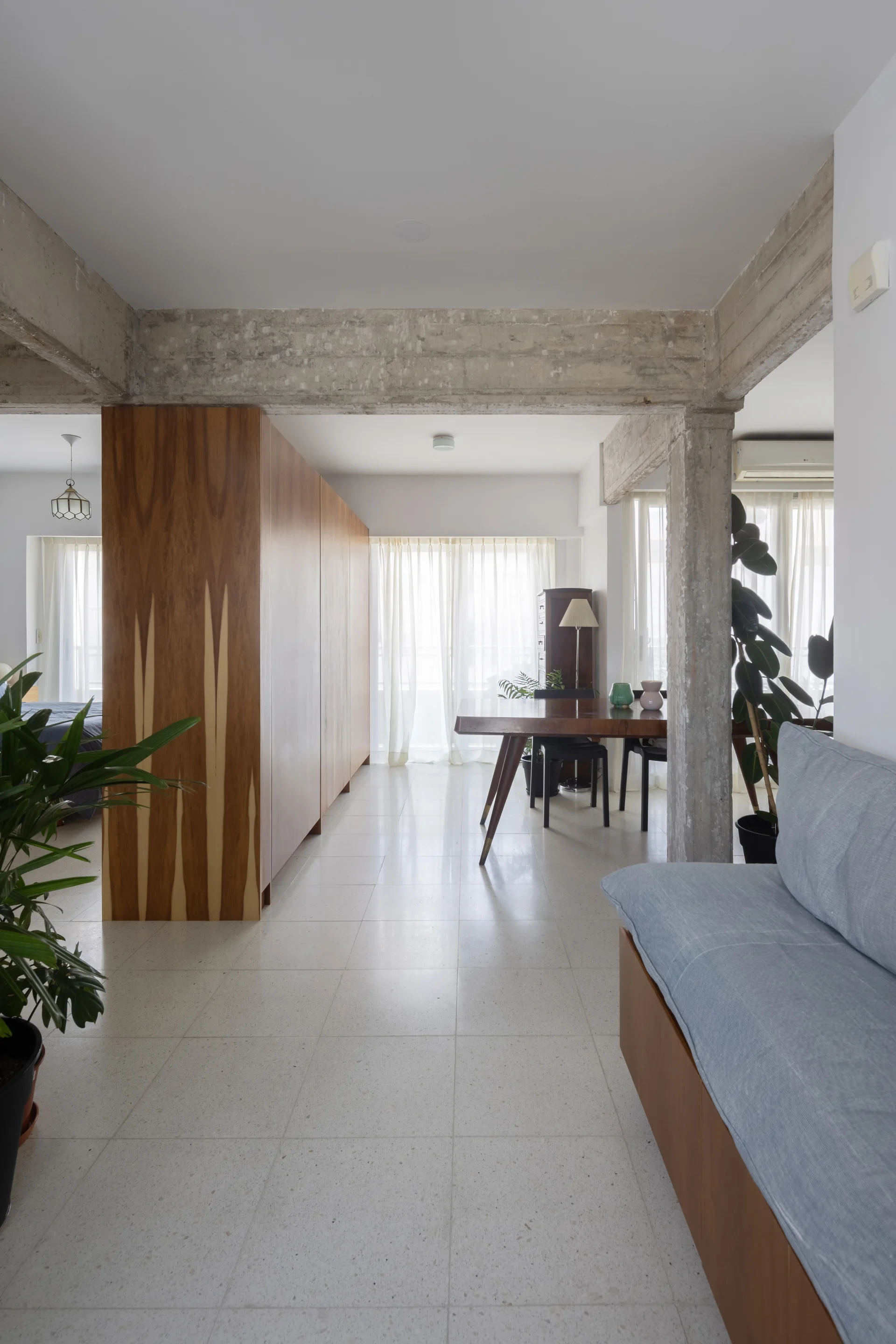 An open apartment interior featuring a large wood cabinet room divider, raw concrete ceiling beams, and a dining table set near sheer curtains.