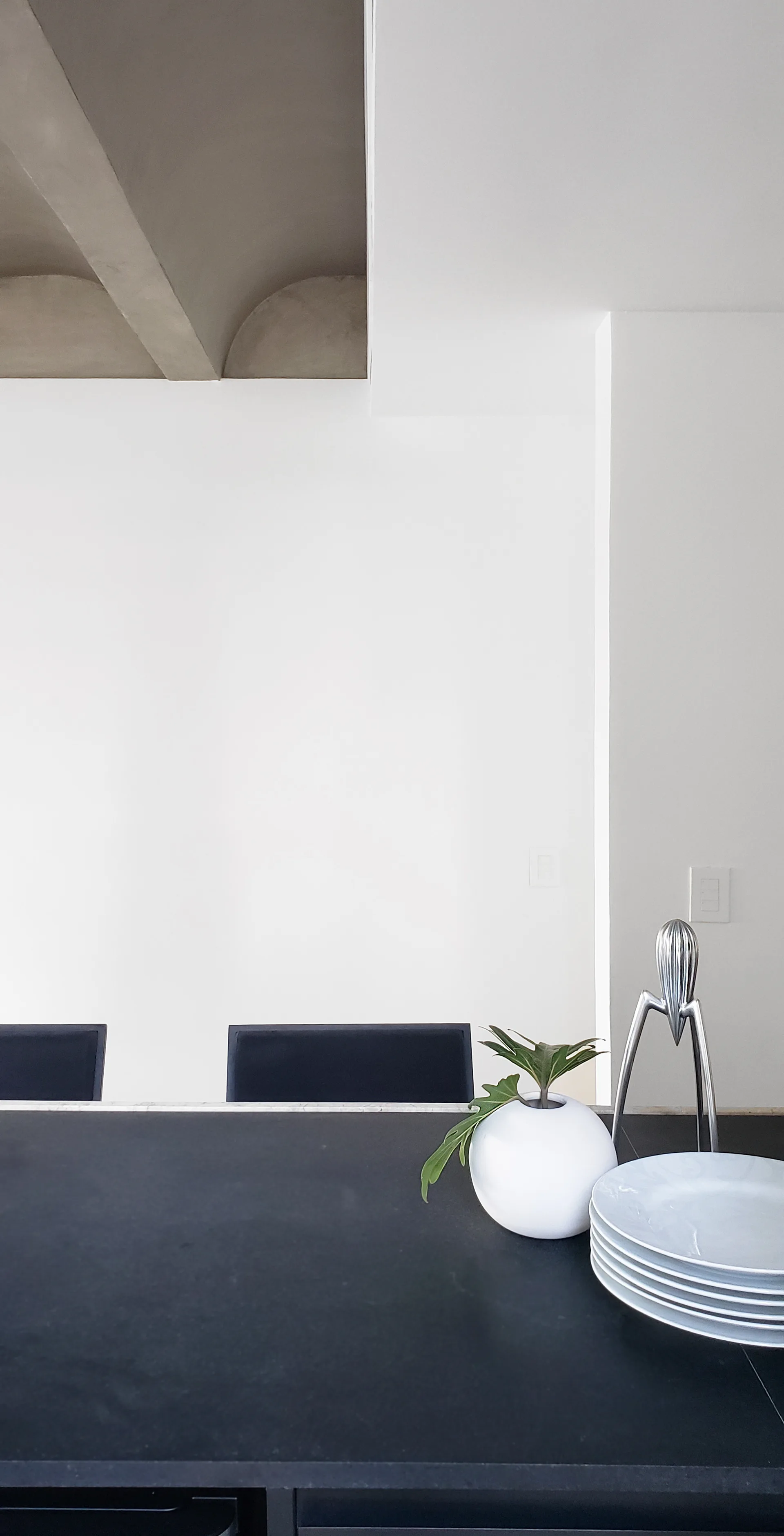 A minimalist kitchen island with a dark matte countertop features a sculptural metal juicer, a stack of white plates, and a concrete ceiling structure above.