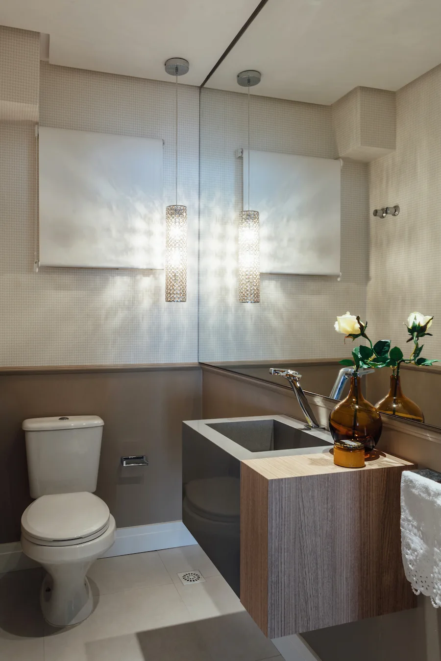 A modern powder room features a white toilet, a wall-mounted wood and grey vanity, and a cylindrical crystal pendant light reflecting in a large mirror.