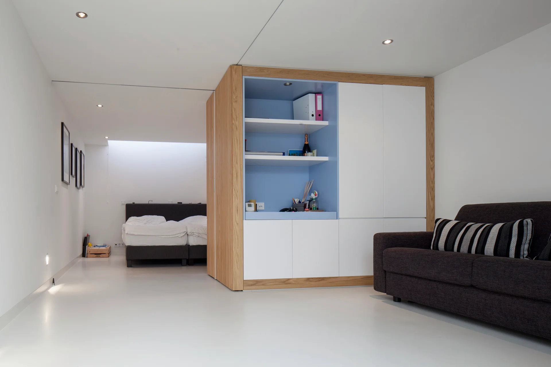 A custom wood and white storage cabinet acts as a room partition between a modern living area with a dark sofa and a recessed bedroom.
