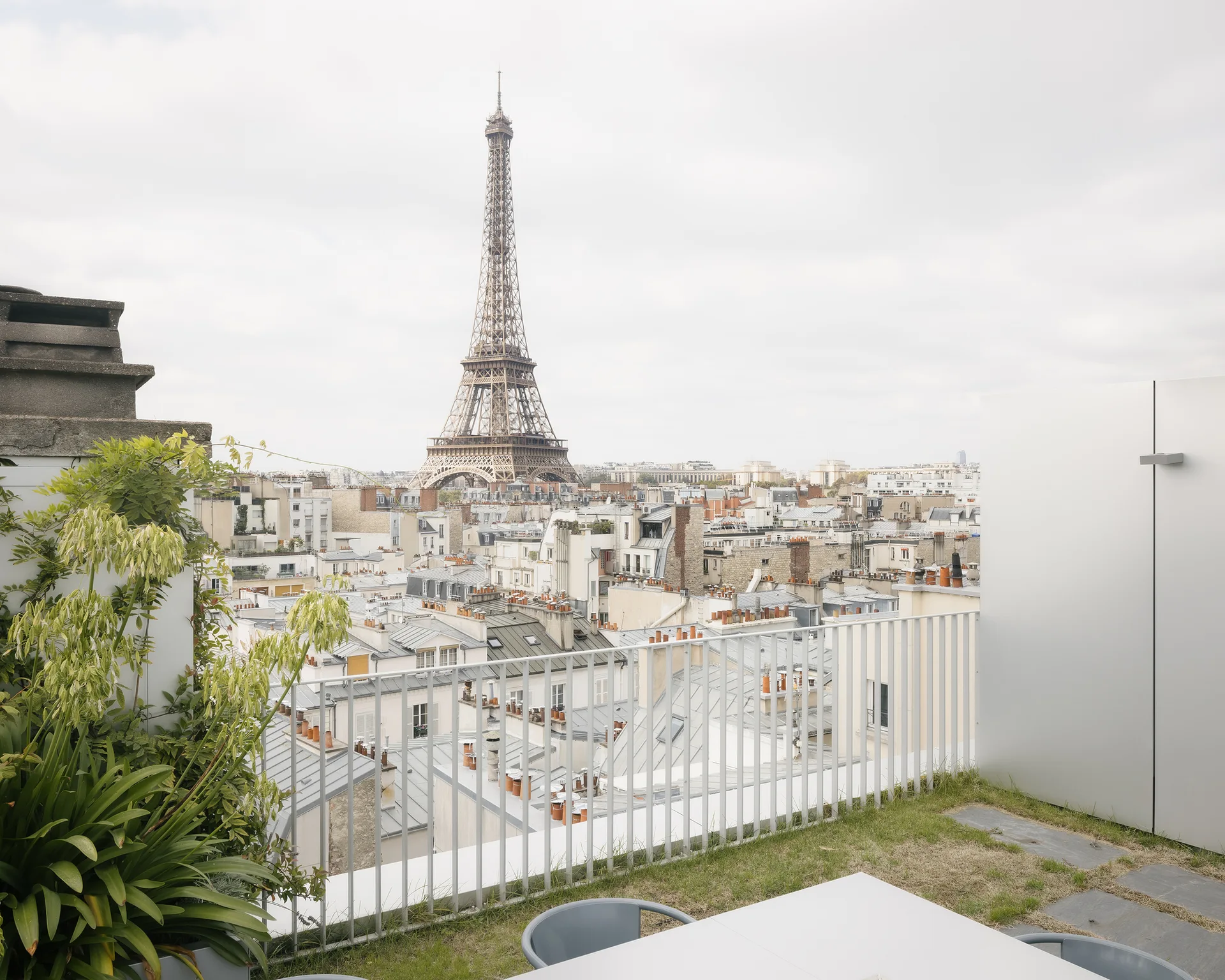 An elevated rooftop terrace with a white dining table, metal railing, and a clear view of the Eiffel Tower over Parisian rooftops.