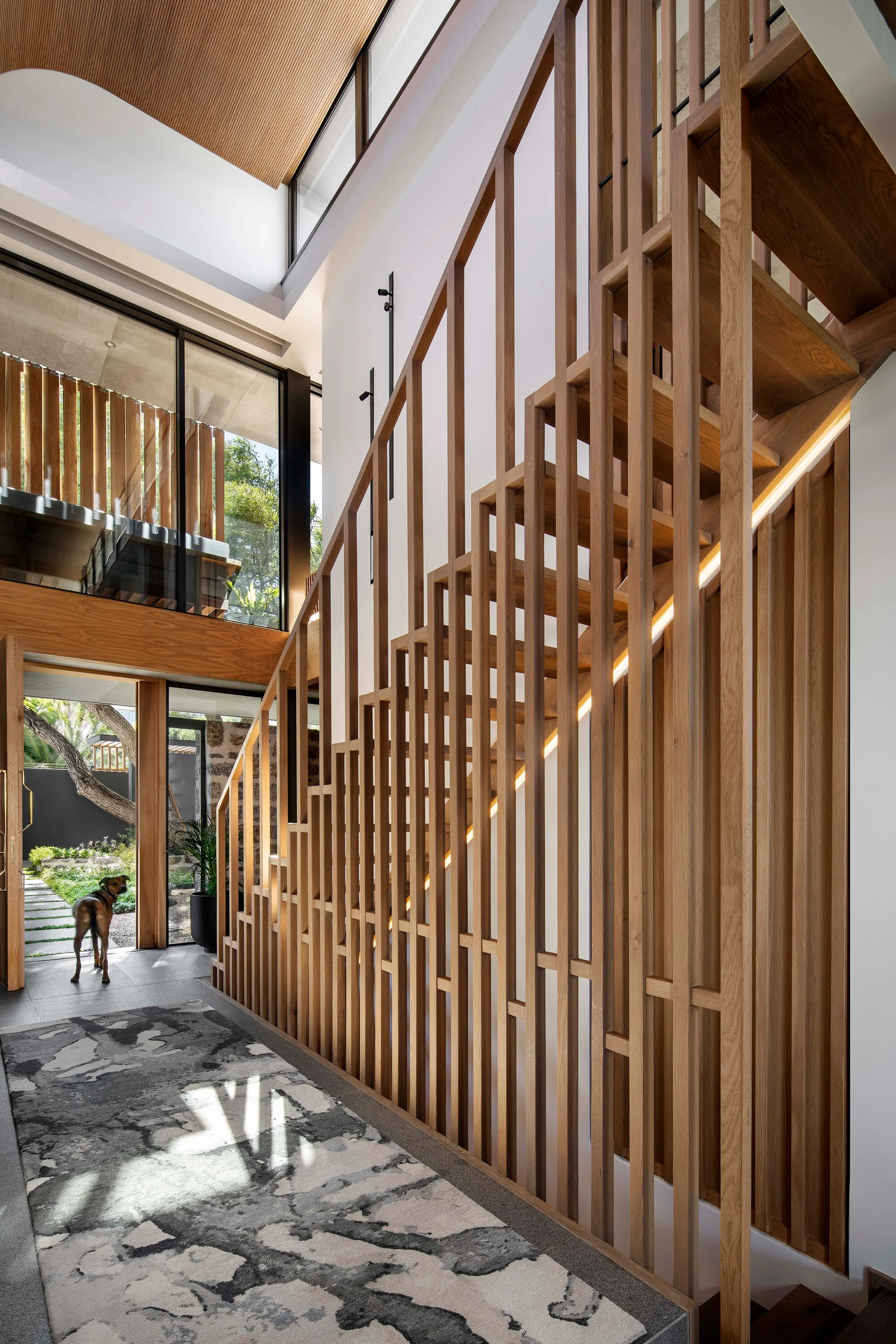 A modern hallway featuring a staircase with a vertical wood-slat balustrade, integrated LED light strip, and patterned rug.