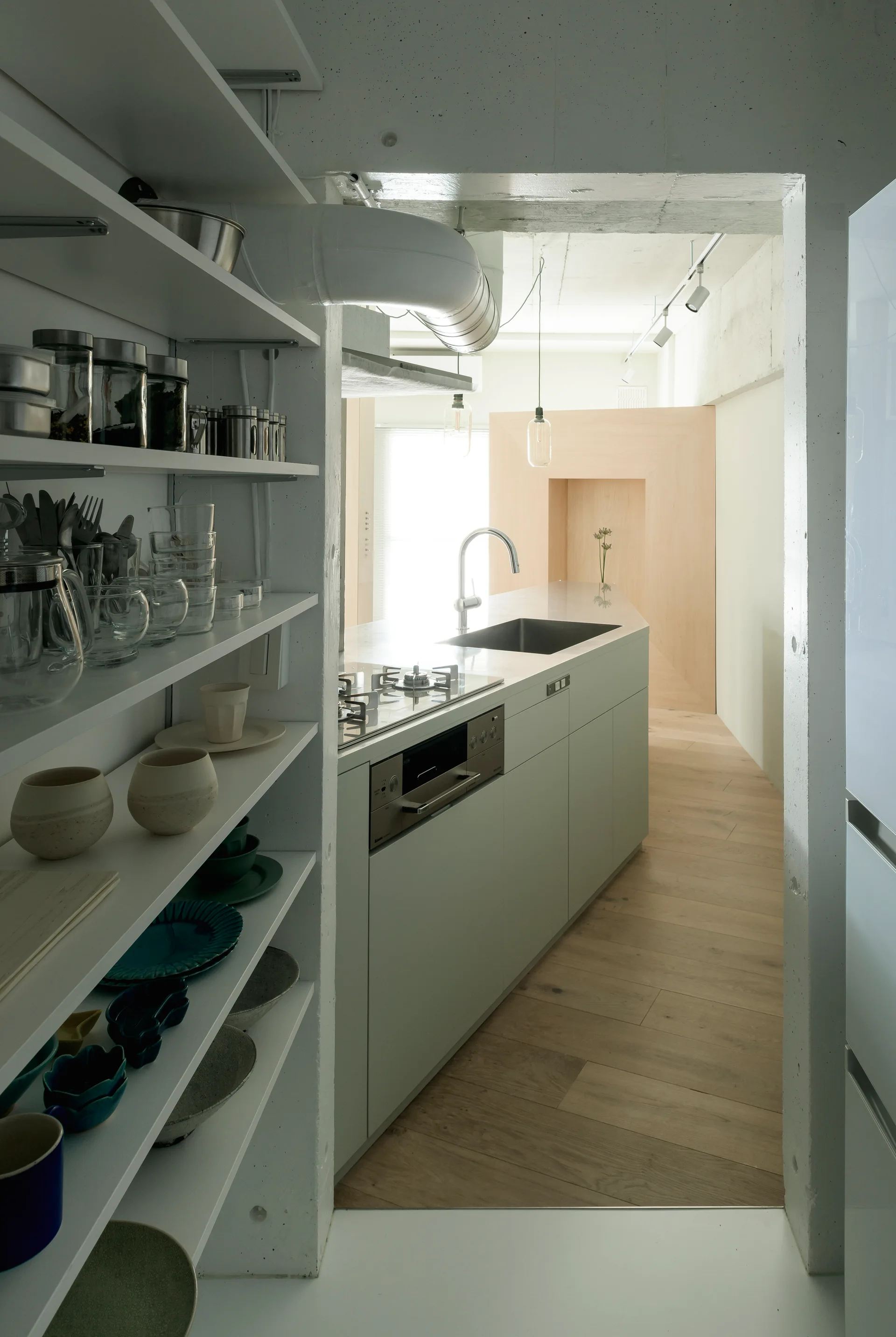 A modern kitchen viewed through a concrete entryway features a light grey island with a sink and open shelves filled with dishes.