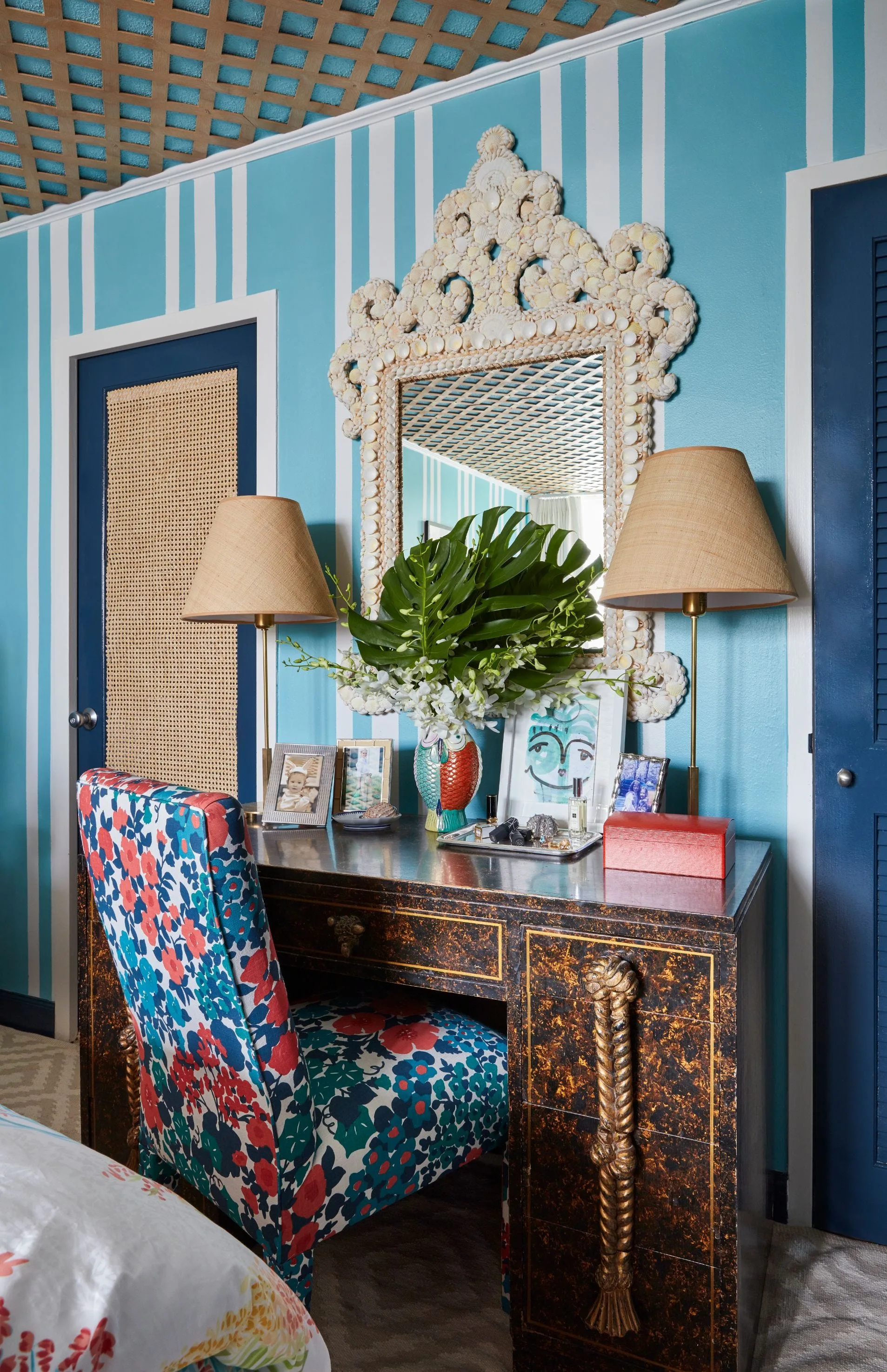 A bedroom writing desk with a floral chair is positioned against turquoise striped walls beneath an ornate shell-framed mirror.
