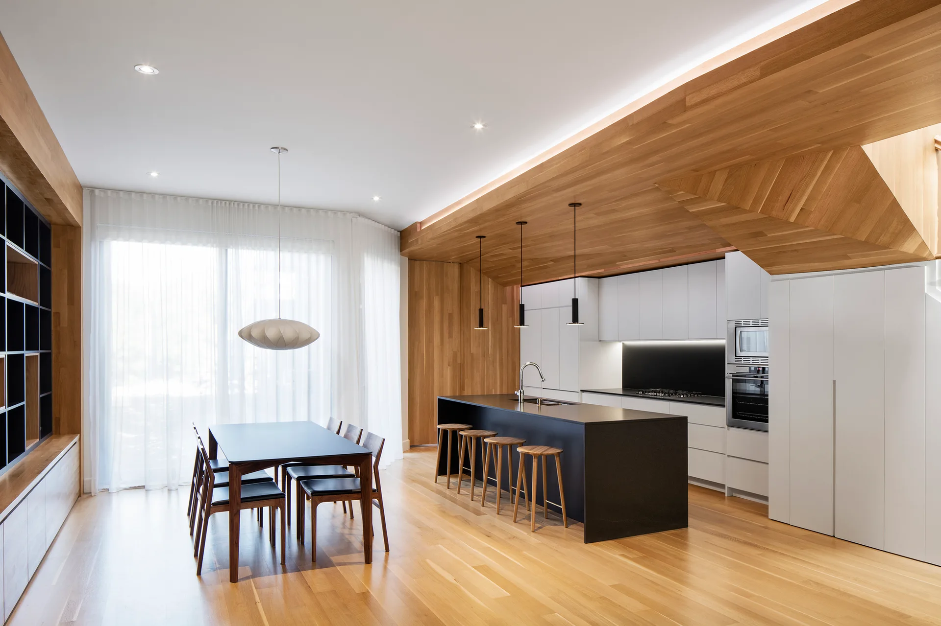 A modern kitchen and dining space with an angled wood ceiling canopy, white cabinetry, a black island, and sheer curtains.