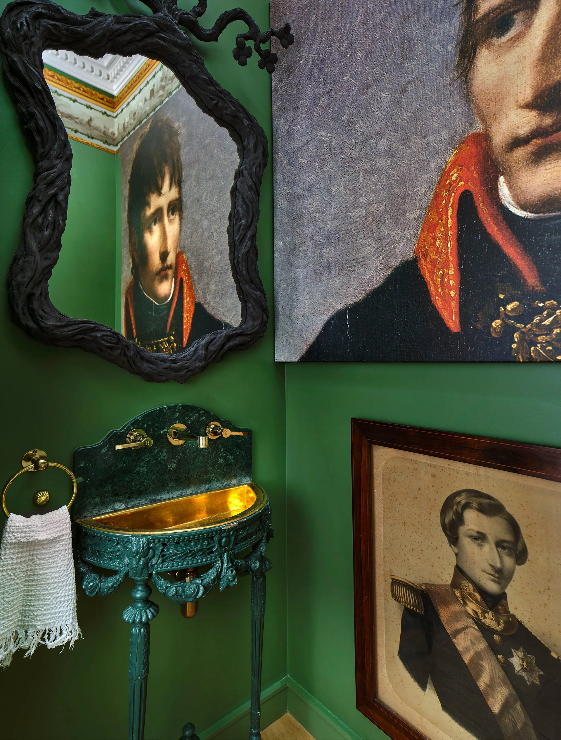 An eclectic powder room with forest green walls, an ornate pedestal sink featuring a gold basin, a textured black mirror, and framed vintage portrait art.