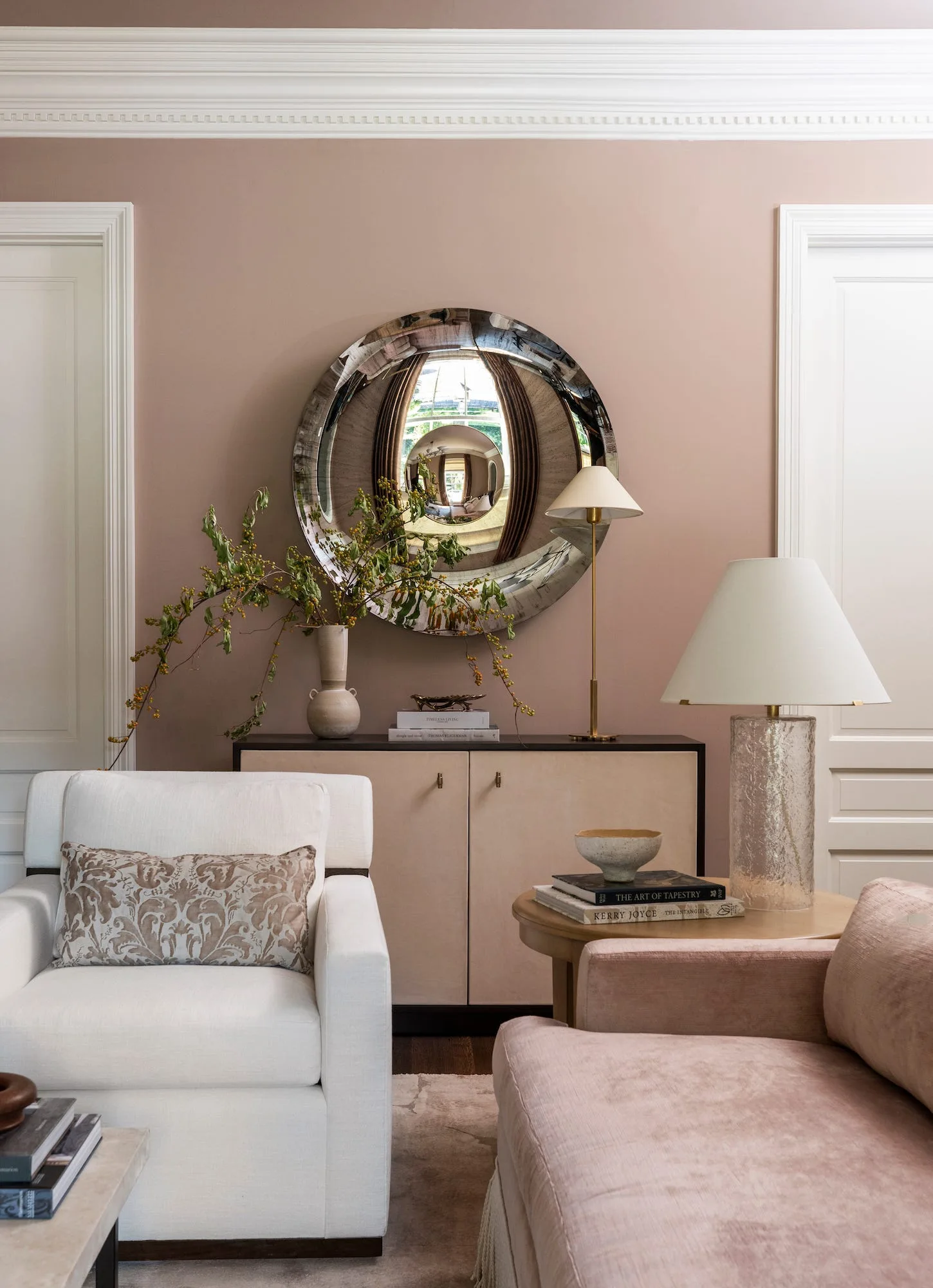 A large round convex mirror hangs on a mauve wall above a beige sideboard flanked by white doors, an armchair, and a side table.