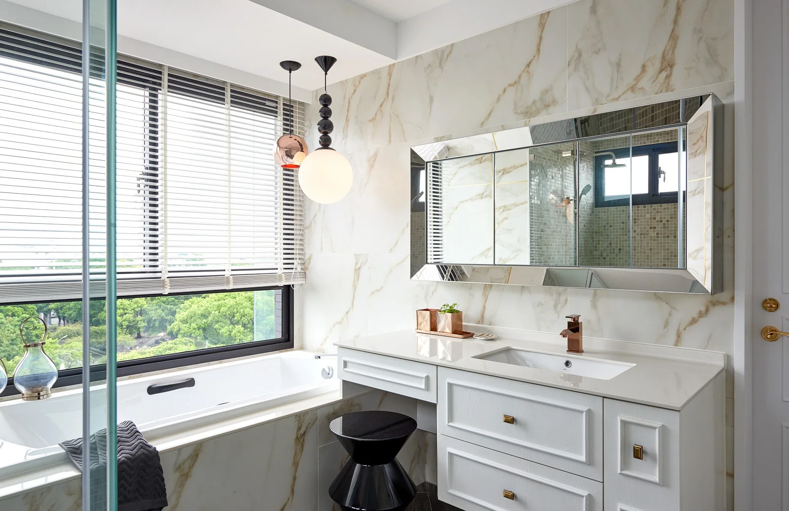 A modern bathroom features marble-look tiled walls, a white vanity with gold hardware, a large framed mirror, and a bathtub under a window.