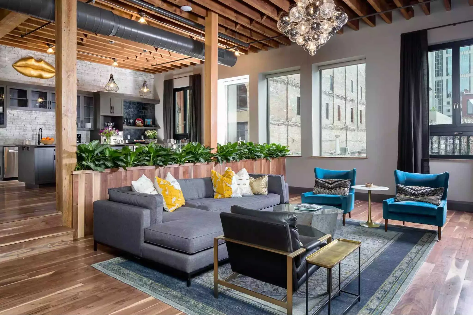 An industrial loft living room with a grey sectional, blue velvet armchairs, and a wood partition wall topped with leafy green plants.