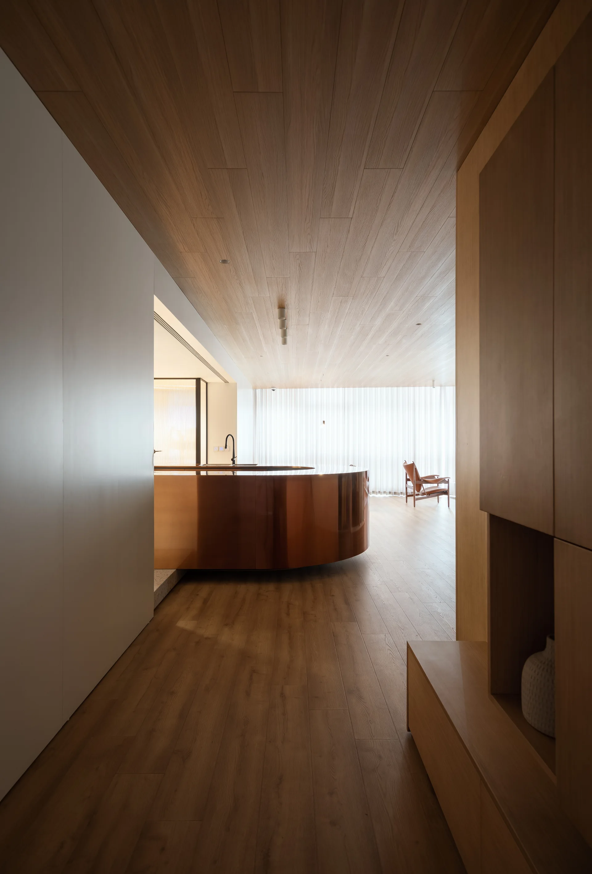 A modern kitchen hallway featuring a curved copper kitchen island, extensive wood paneling on the floor and ceiling, and a sheer curtain.
