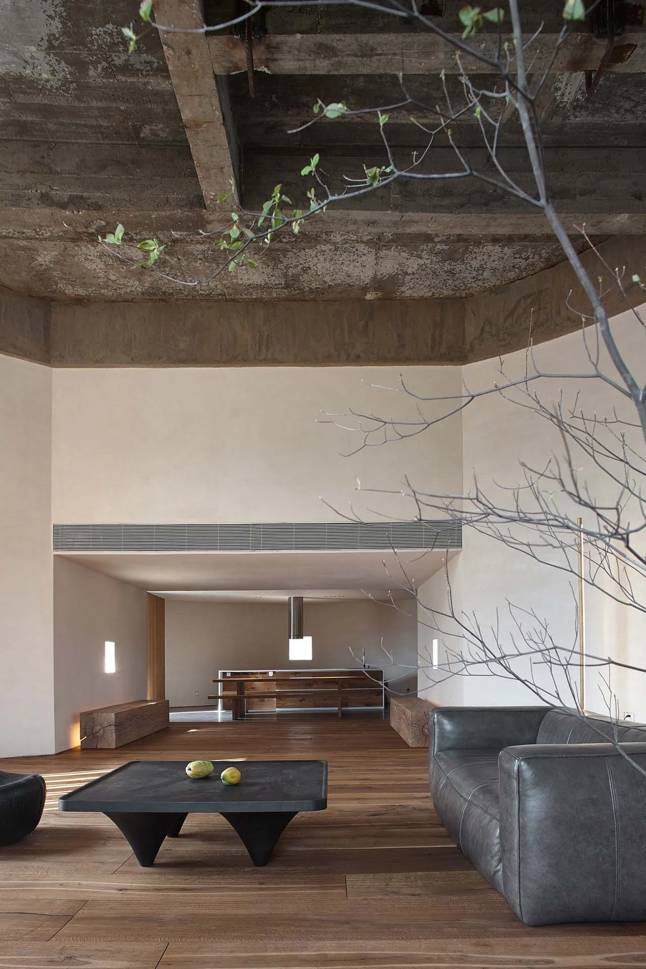 A minimalist living room features a dark leather sofa, low black coffee table, raw concrete ceiling, and smooth beige plaster walls.