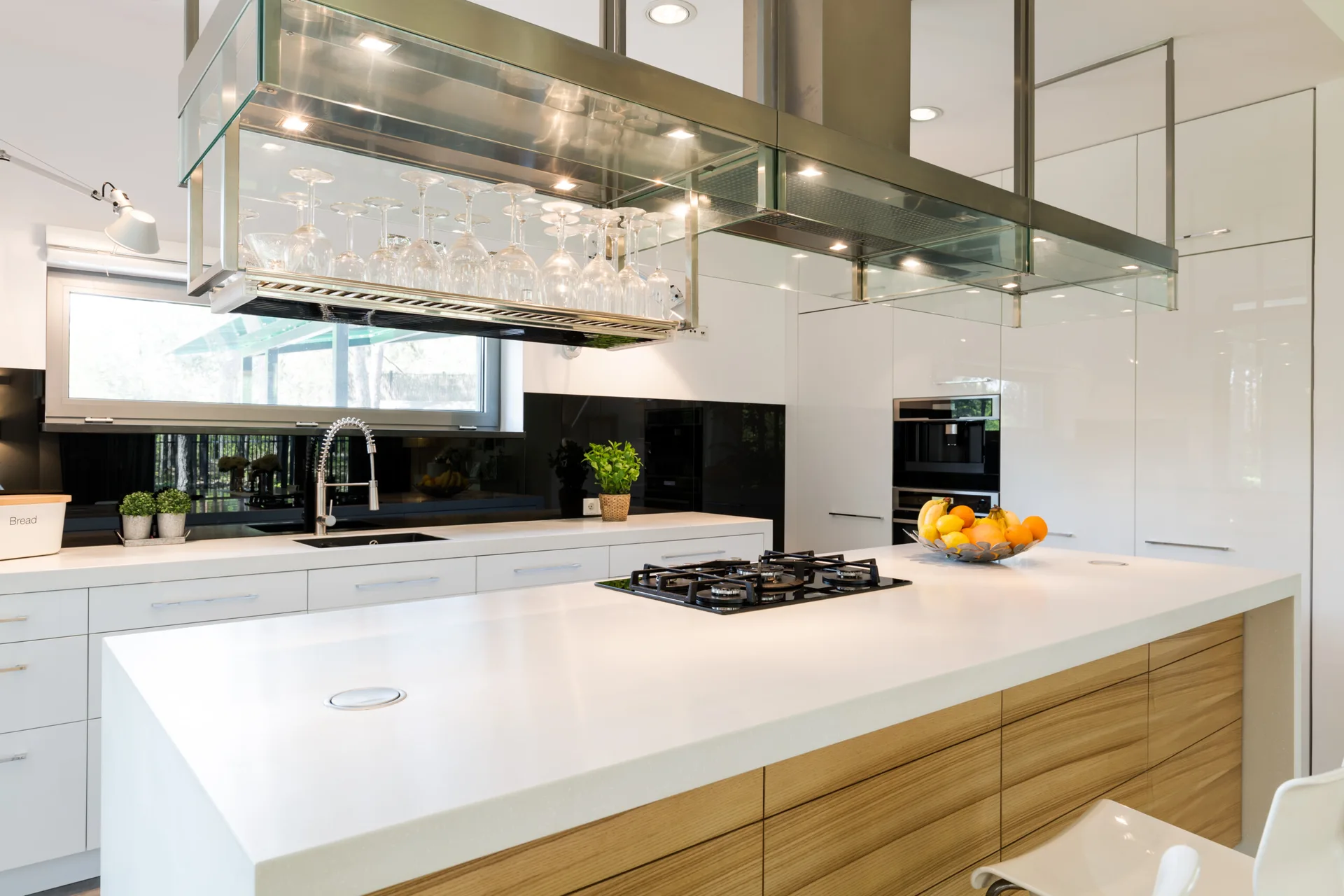 A modern kitchen featuring a white island with a wood-grain base, glossy cabinetry, and a glass-and-metal overhead range hood holding stemware.