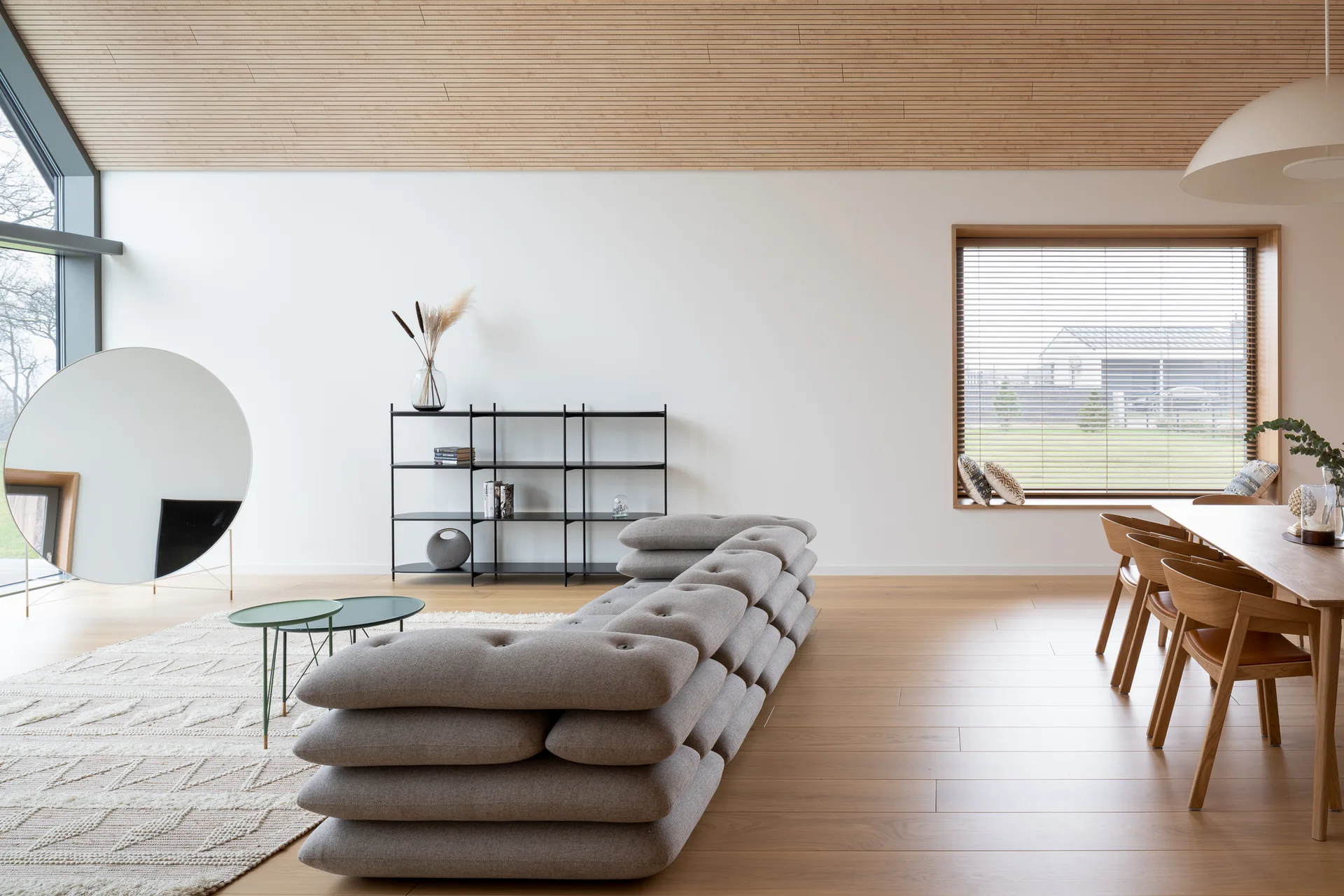 A modern open-concept living and dining area featuring a low-profile grey modular sofa, a slatted wood ceiling, and a large round floor mirror.