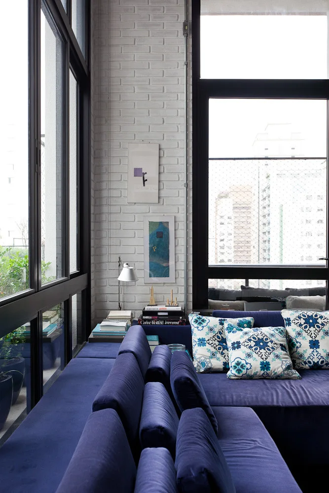 A deep blue sectional sofa sits against a white painted brick wall in a room with tall black-framed windows.