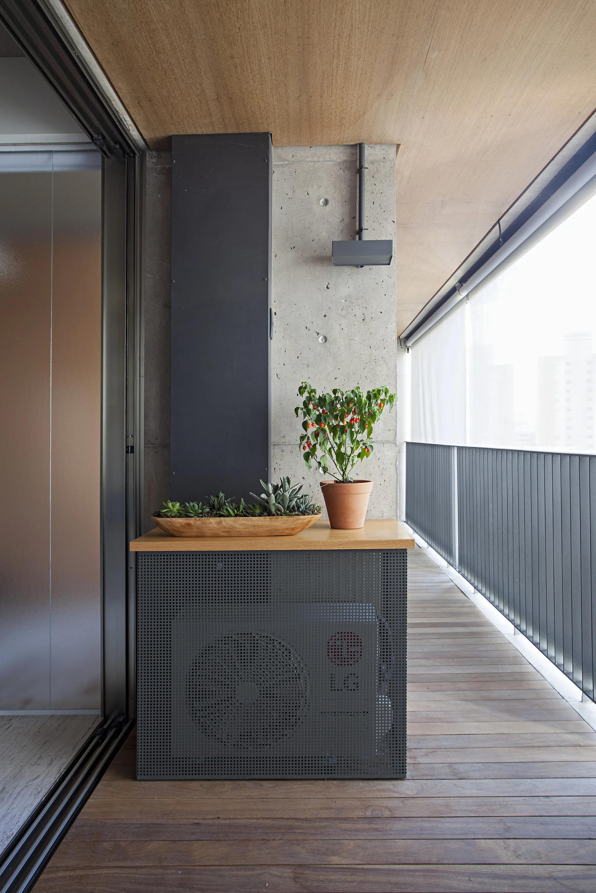 An outdoor balcony features wood plank flooring, a concrete wall, and a grey perforated metal cabinet topped with potted plants.