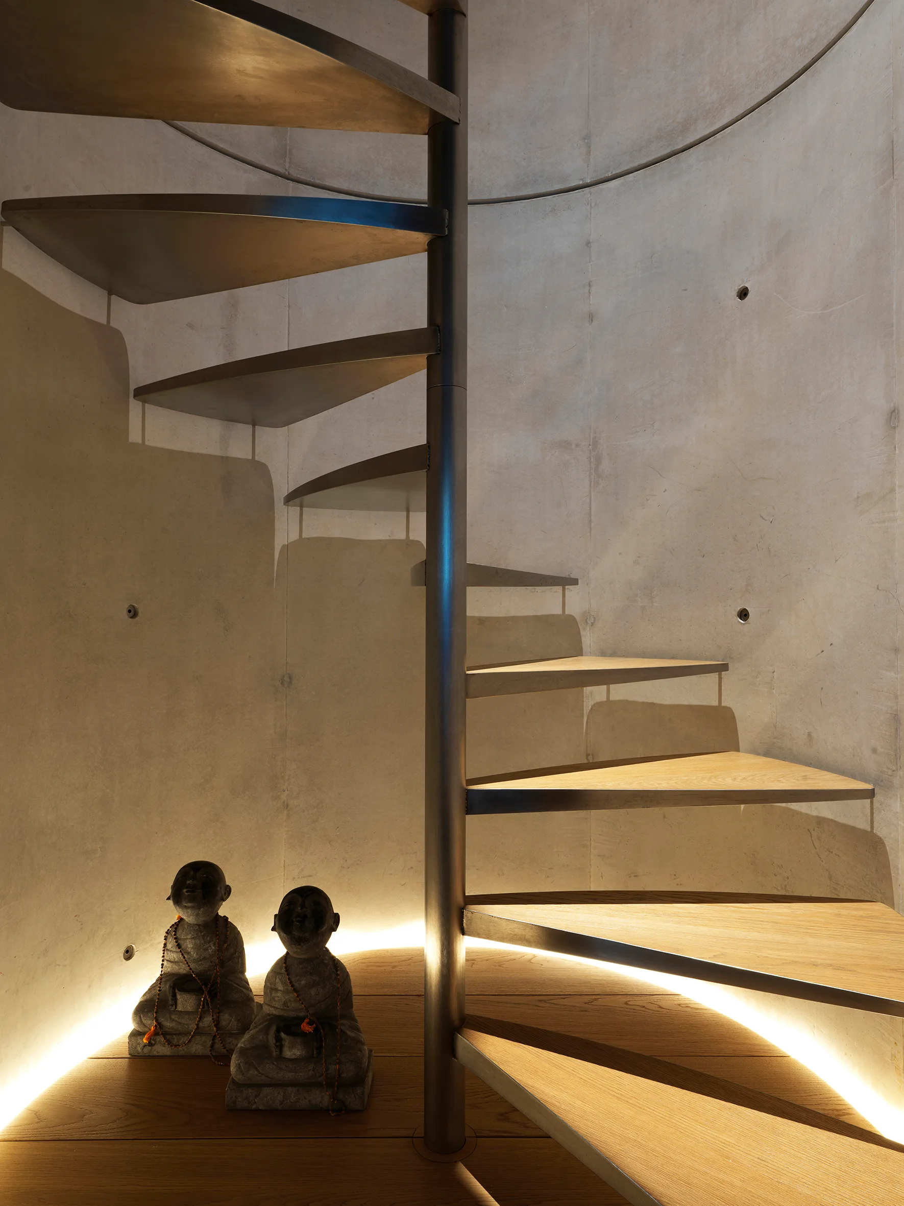 A spiral staircase with wooden steps and a metal spine curves against a smooth concrete wall, lit from below by hidden warm LEDs.