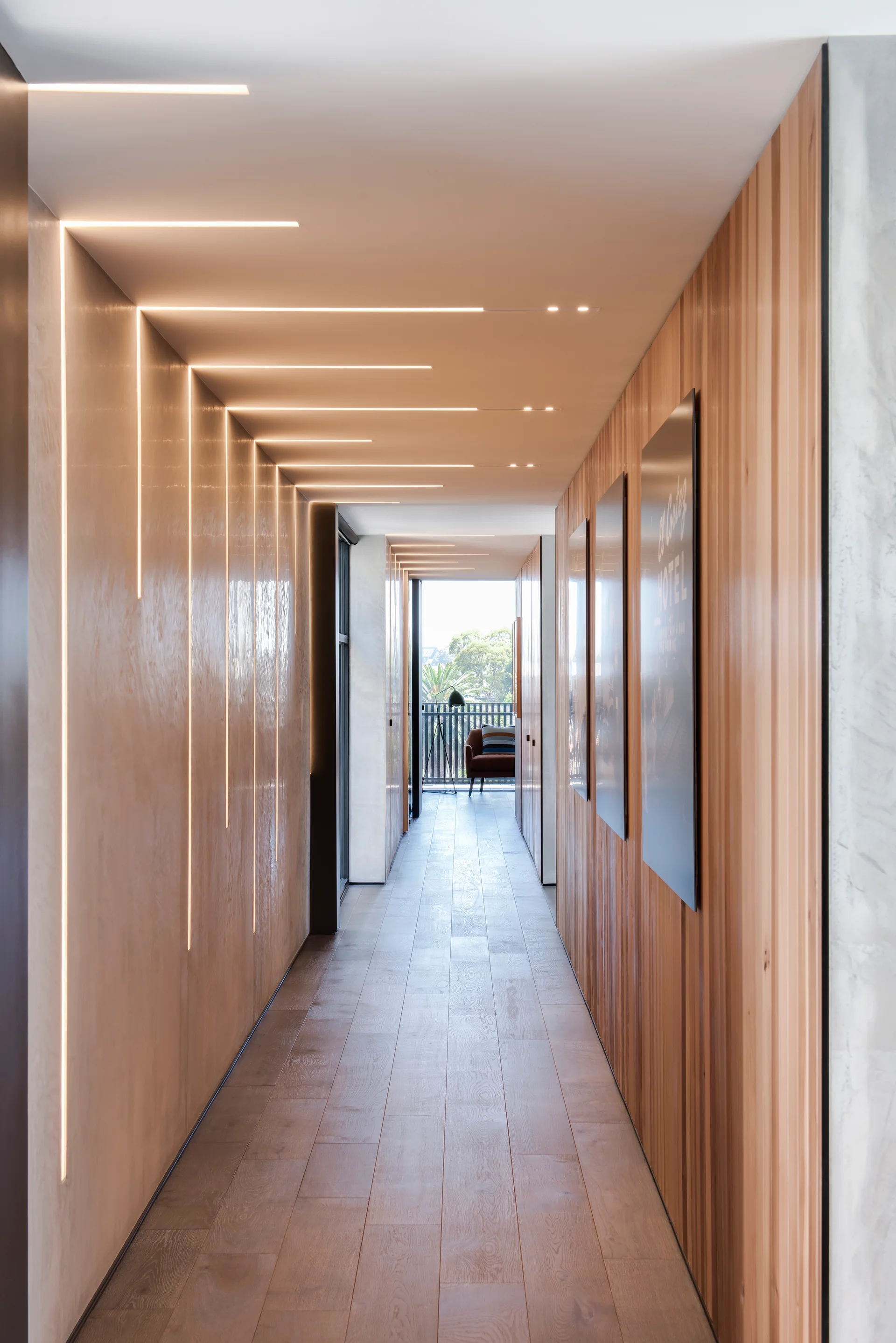 A long modern hallway with integrated linear recessed lighting on the ceiling and walls, featuring light wood slat paneling and hardwood floors.