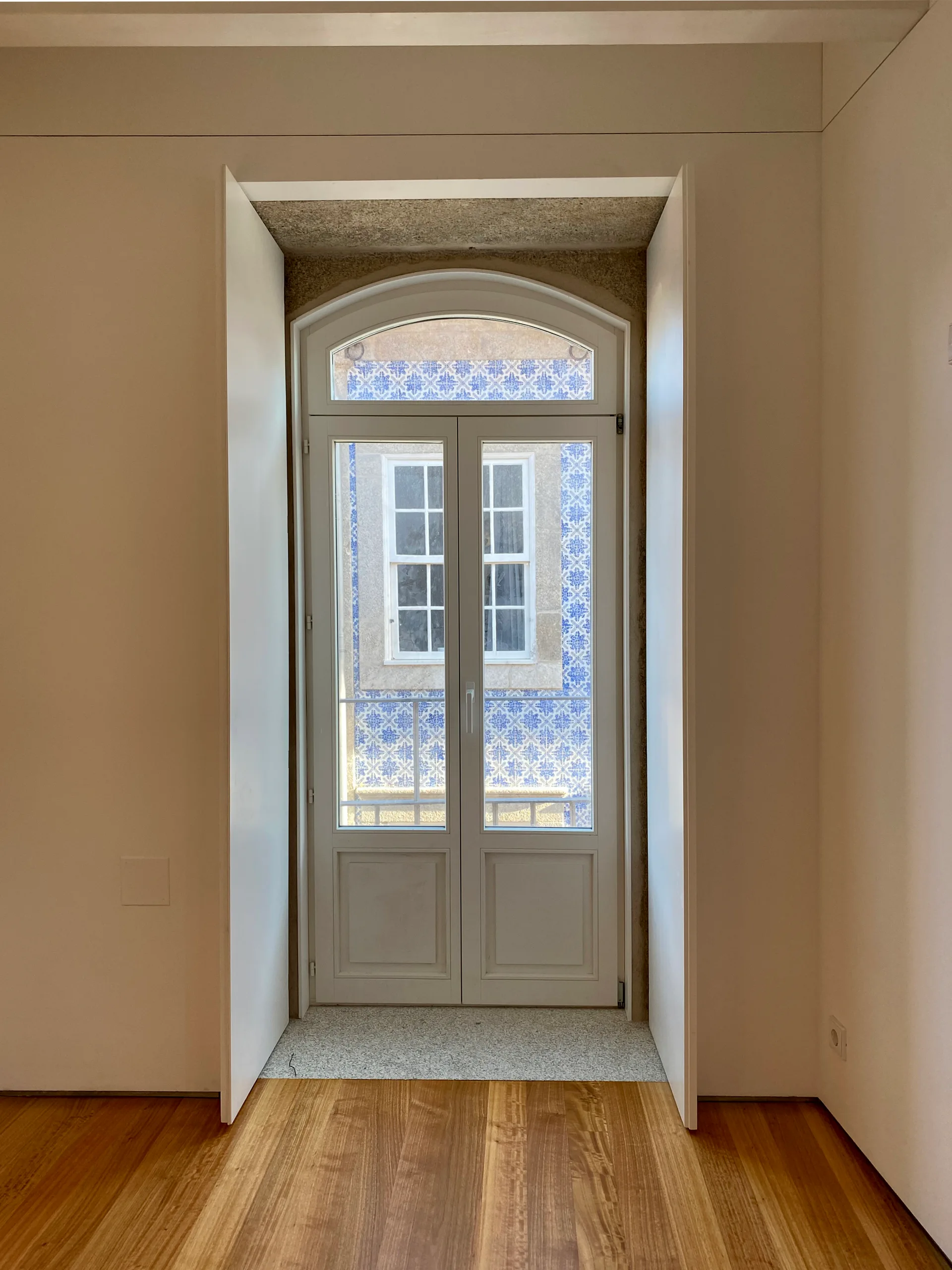A tall arched double door window is recessed into a deep white wall above wood floors, showing a view of blue and white exterior tiles.