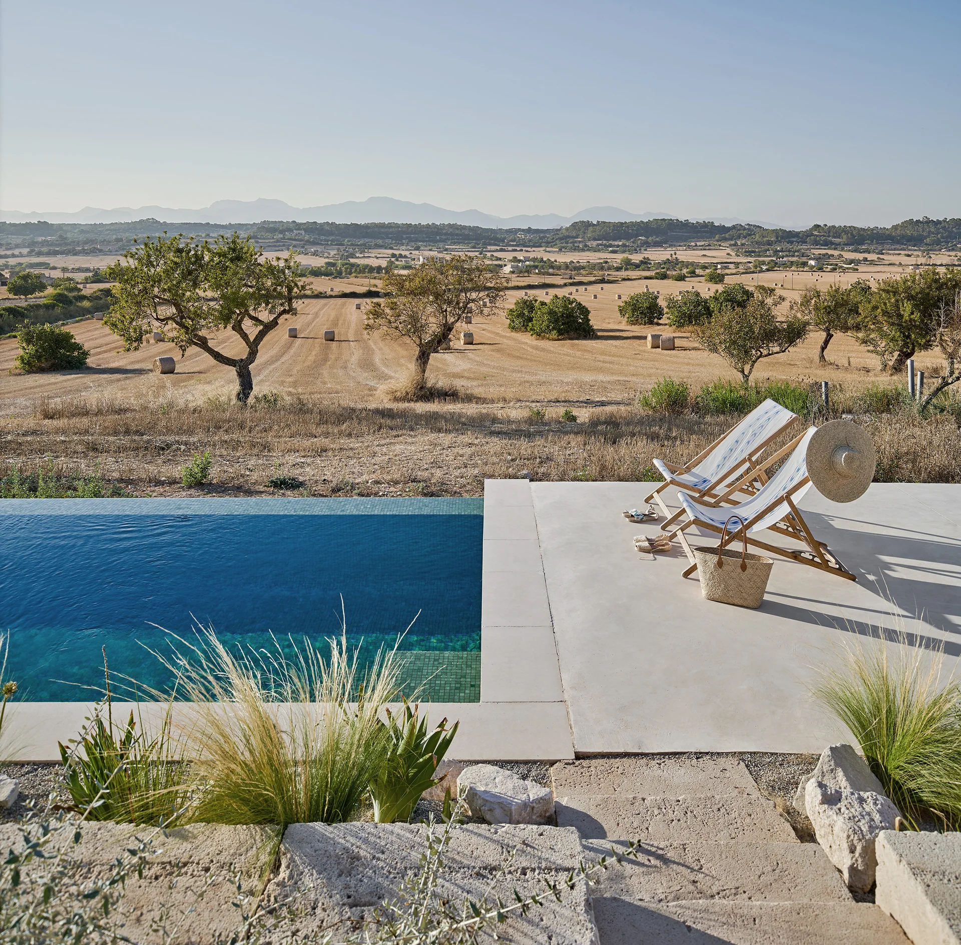 A blue swimming pool sits next to a concrete terrace with two wooden lounge chairs overlooking a rural landscape with mountains.