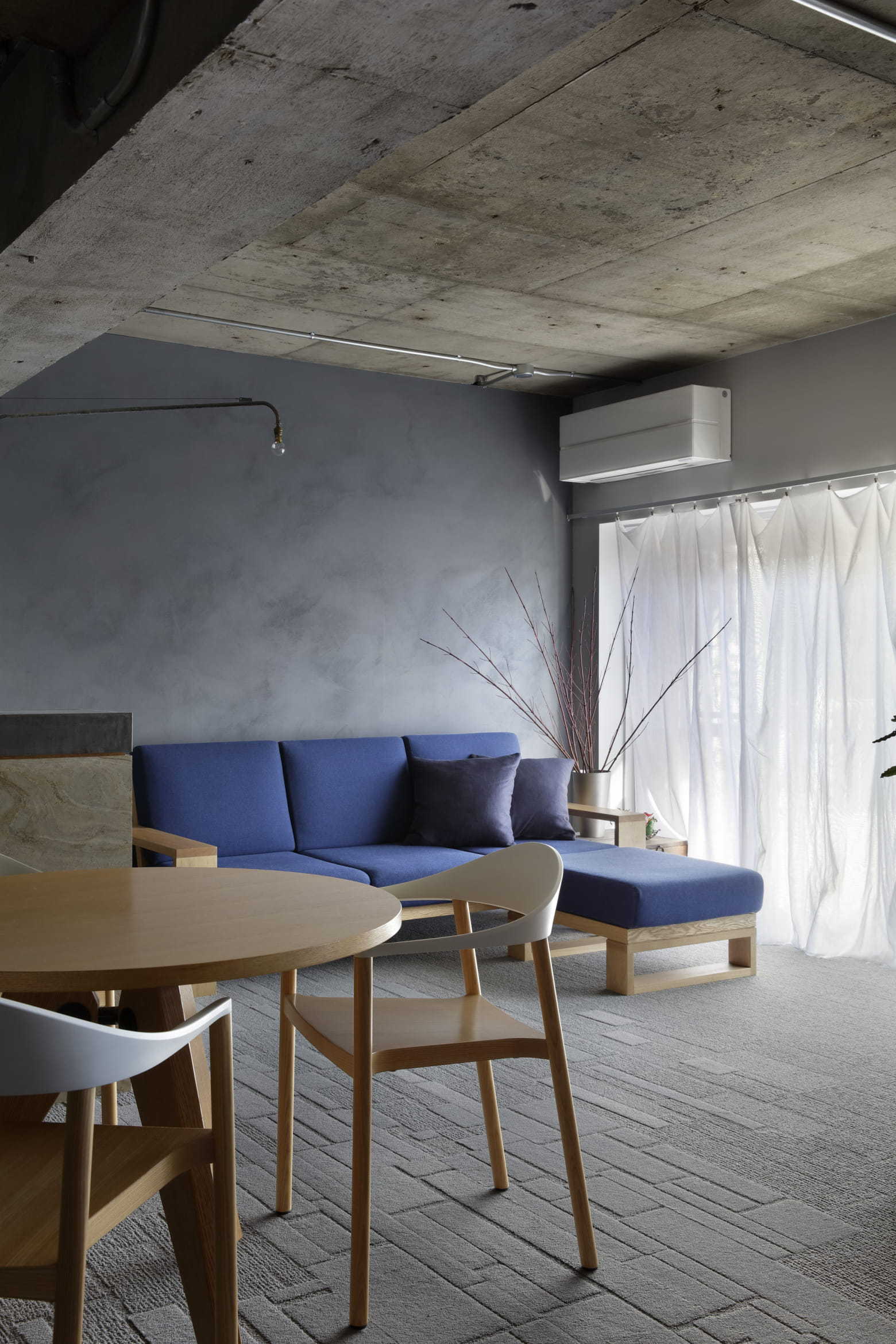 A modern living area features a vibrant blue sofa against a textured gray wall beneath an exposed concrete ceiling.