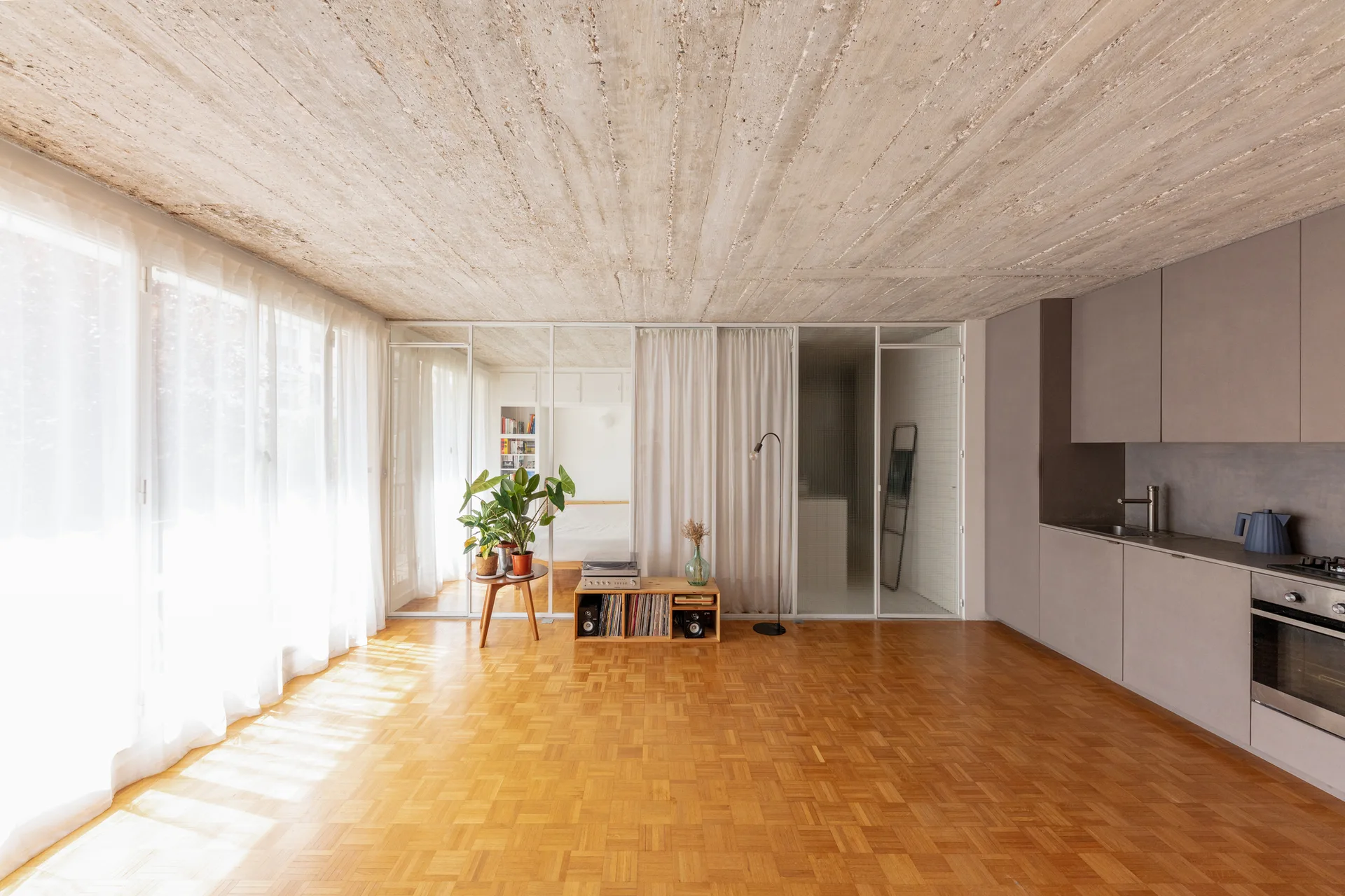 A modern open-plan apartment featuring matte grey kitchen cabinets, a raw board-marked concrete ceiling, and warm patterned parquet flooring.