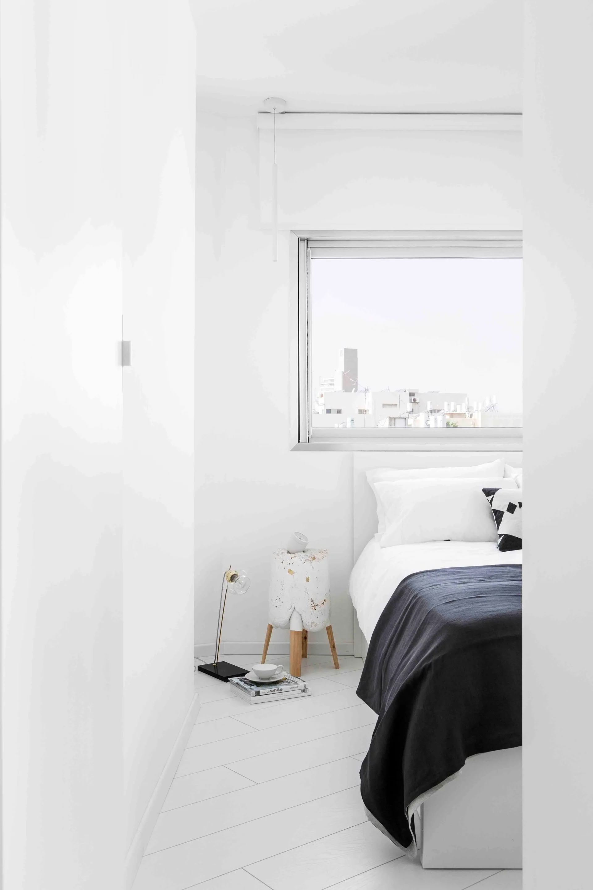 A minimalist white bedroom features a bed with white linens, a dark grey throw, a sculptural wooden-legged side table, and a horizontal window.