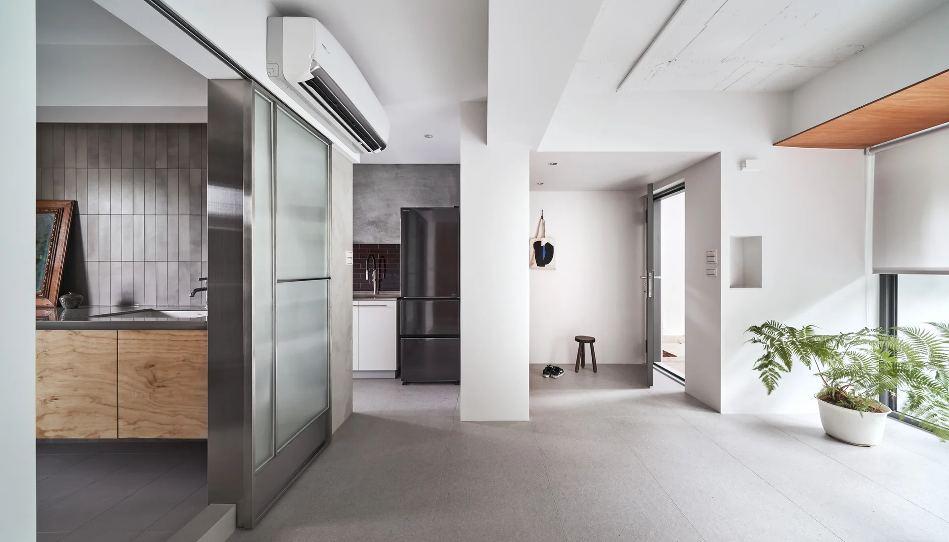 A minimalist entryway with large grey floor tiles, a metal-framed sliding glass door, white walls, and a potted plant by a large window.