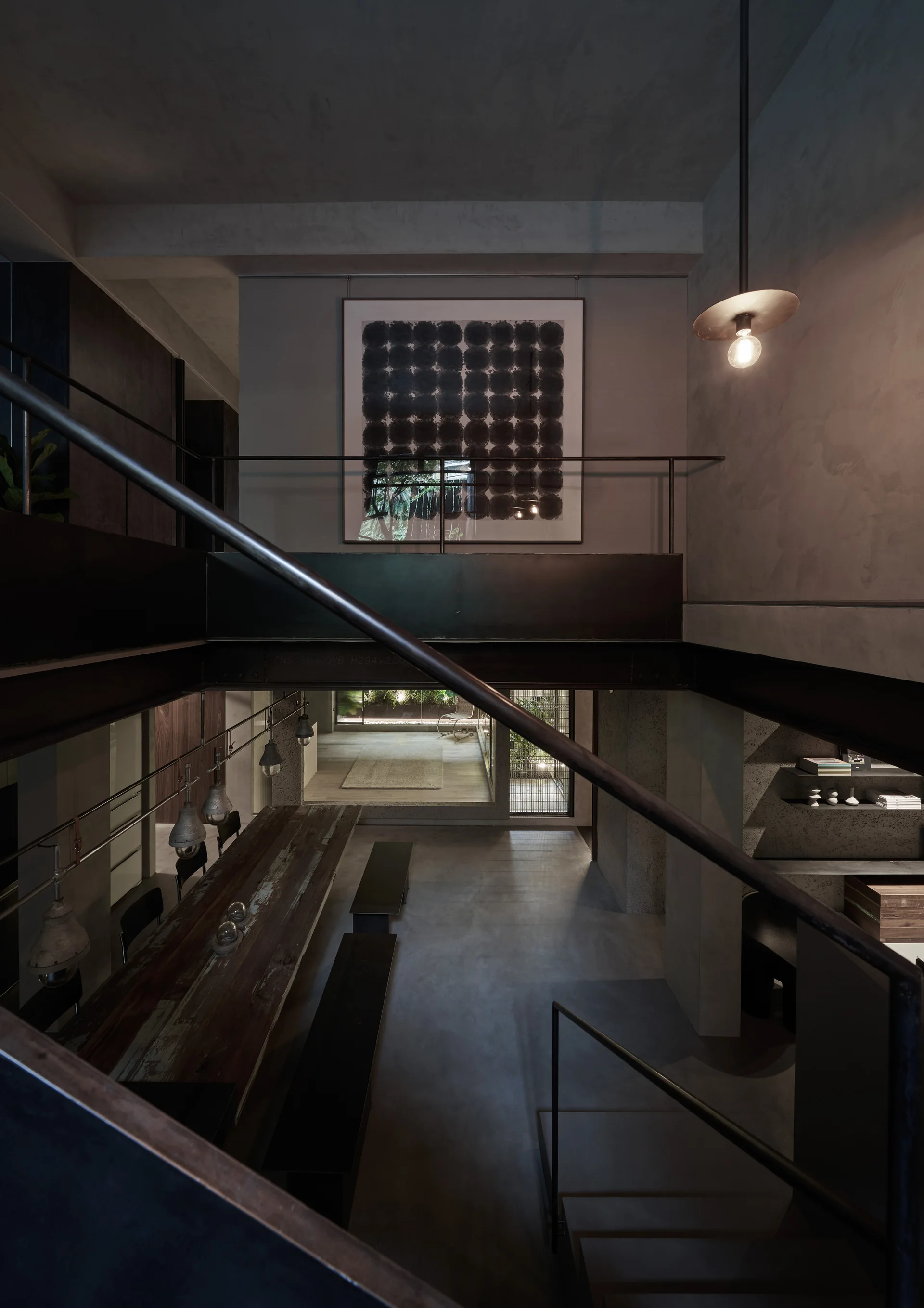 A multi-level industrial dining room features a long weathered wood table, dark benches, and raw concrete walls framed by black metal railings.