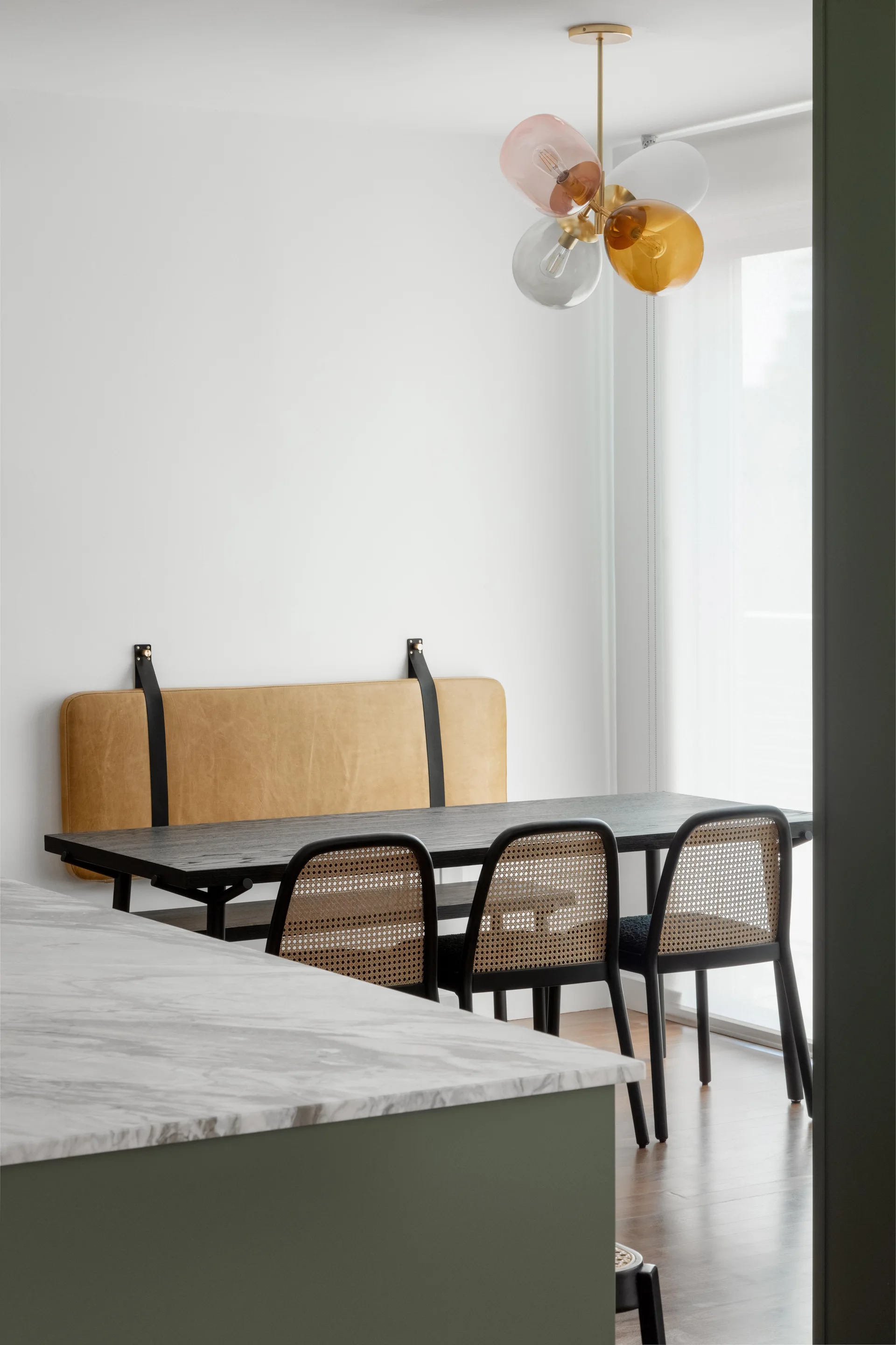 A dining table with cane-back chairs and a wall-mounted leather cushion sits next to a green kitchen island with a marble top.