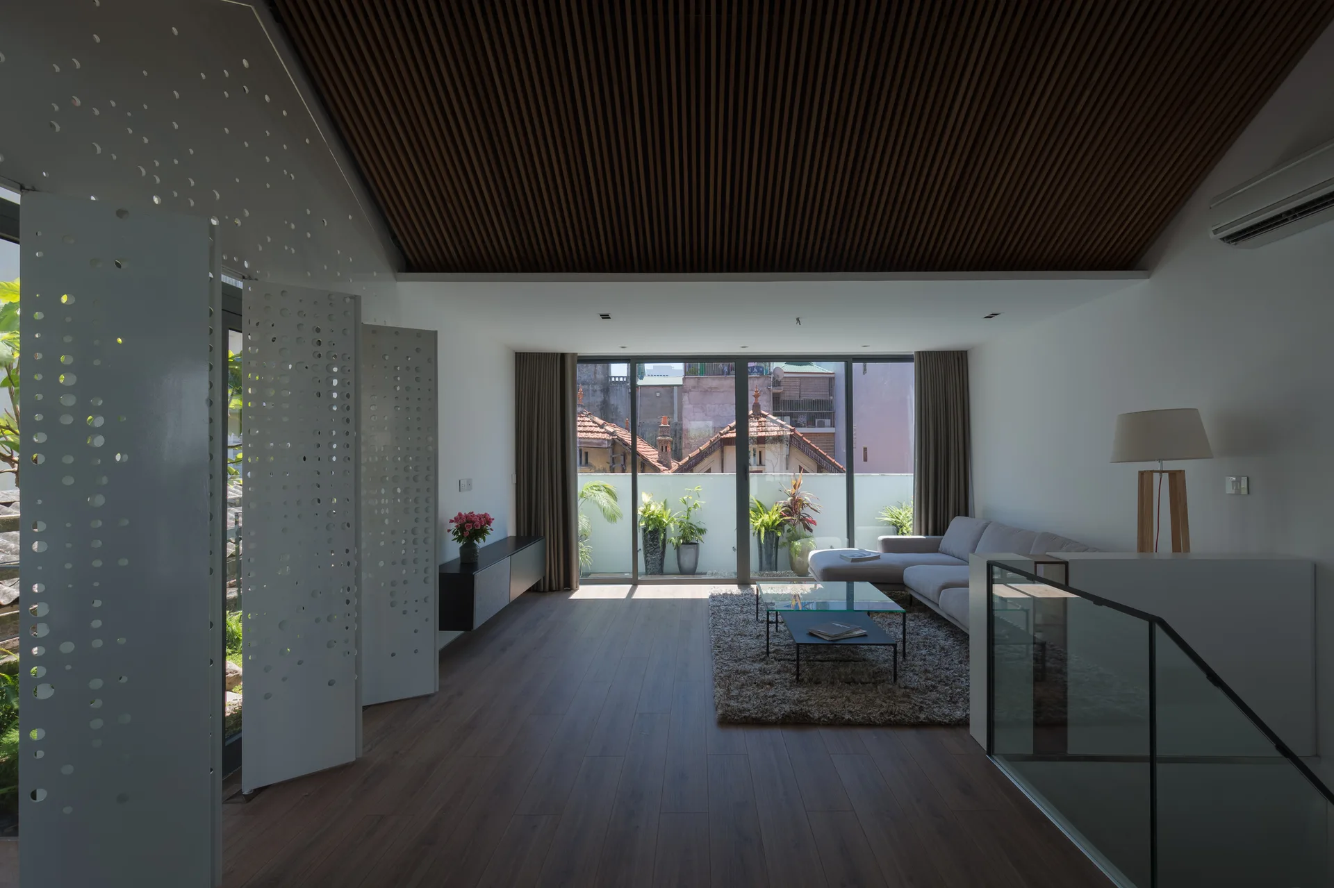 A minimalist living room with a dark wood slatted ceiling, a grey sectional sofa, and sliding glass doors opening to a terrace.